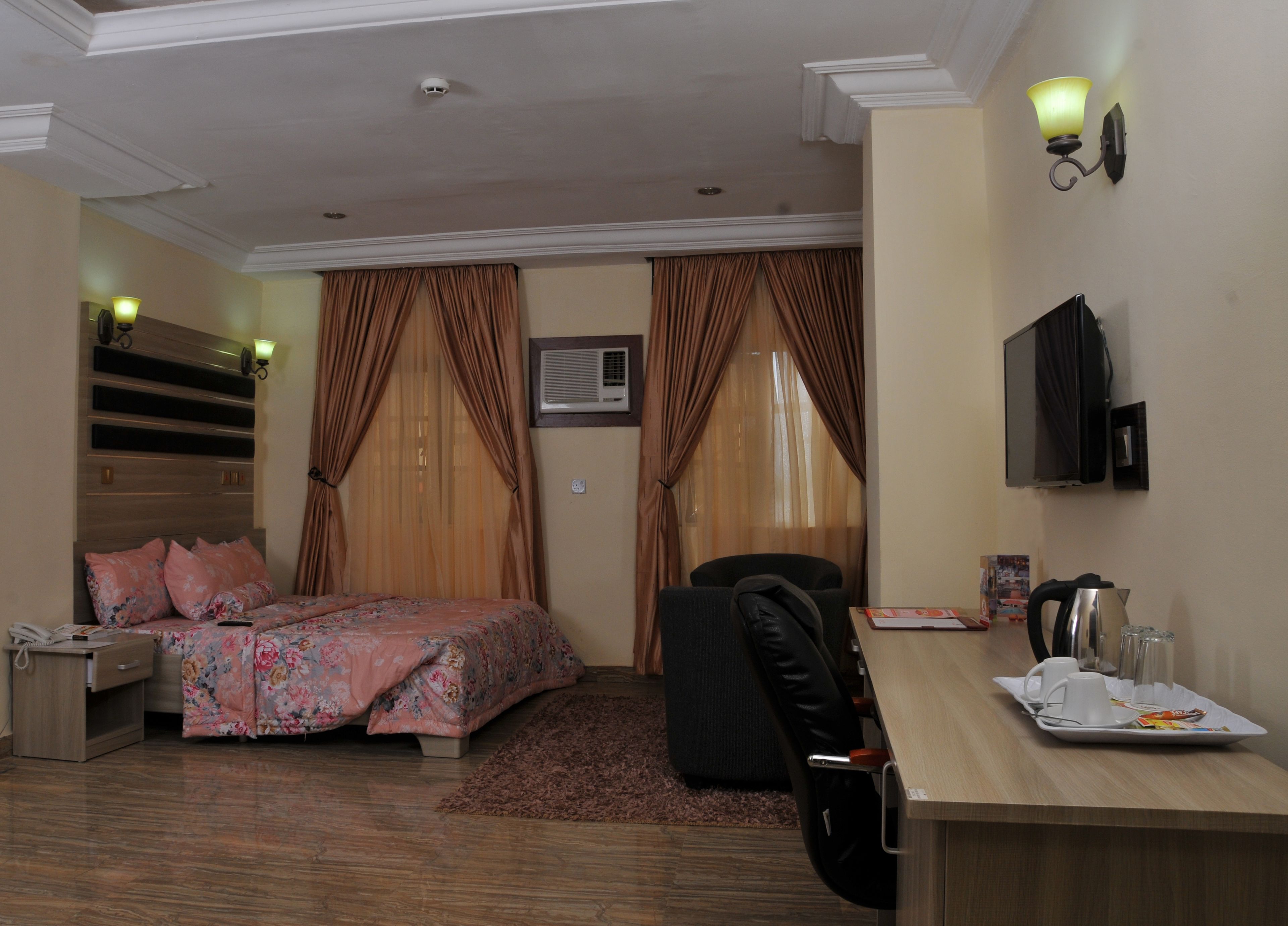 A hotel room with a bed, a desk with a tray of breakfast items, a chair, a TV, and an air conditioning unit.