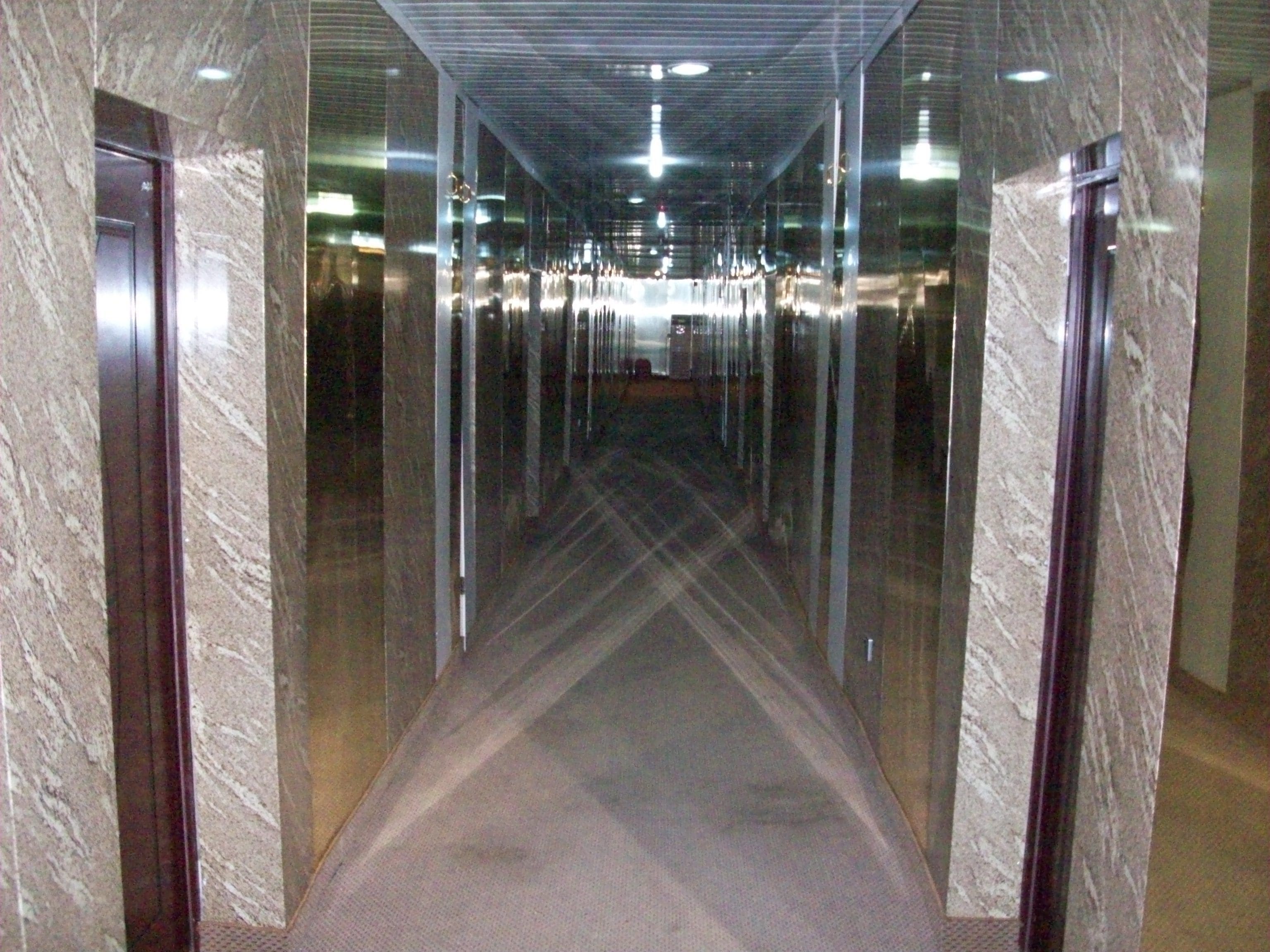 A corridor with glass walls and a carpeted floor, leading to a door at the end.