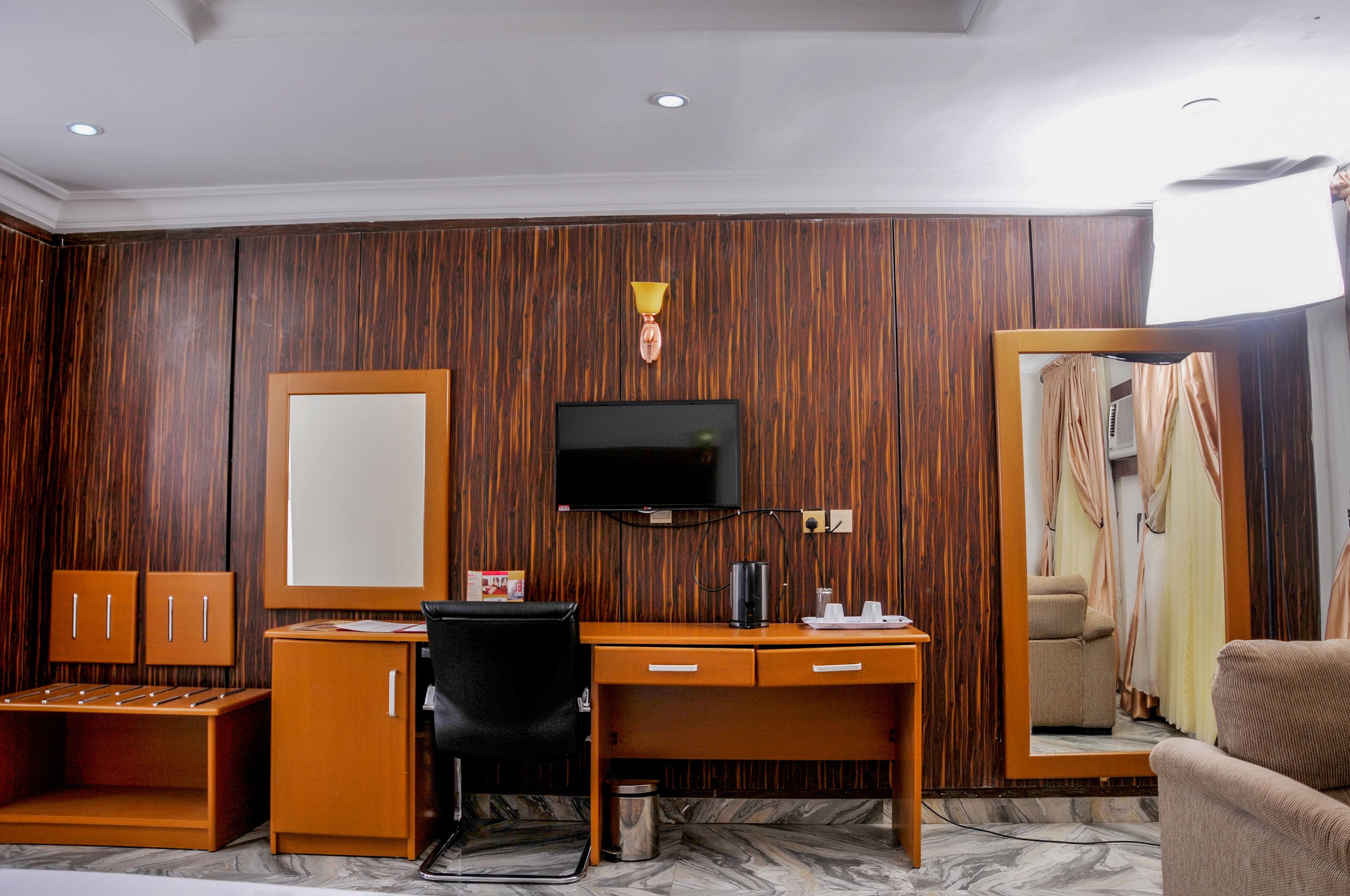 A hotel room with a wooden paneled wall, a flat-screen TV mounted above a wooden desk, a wardrobe with two doors, a black office chair, and a large mirror reflecting a bed and curtains.