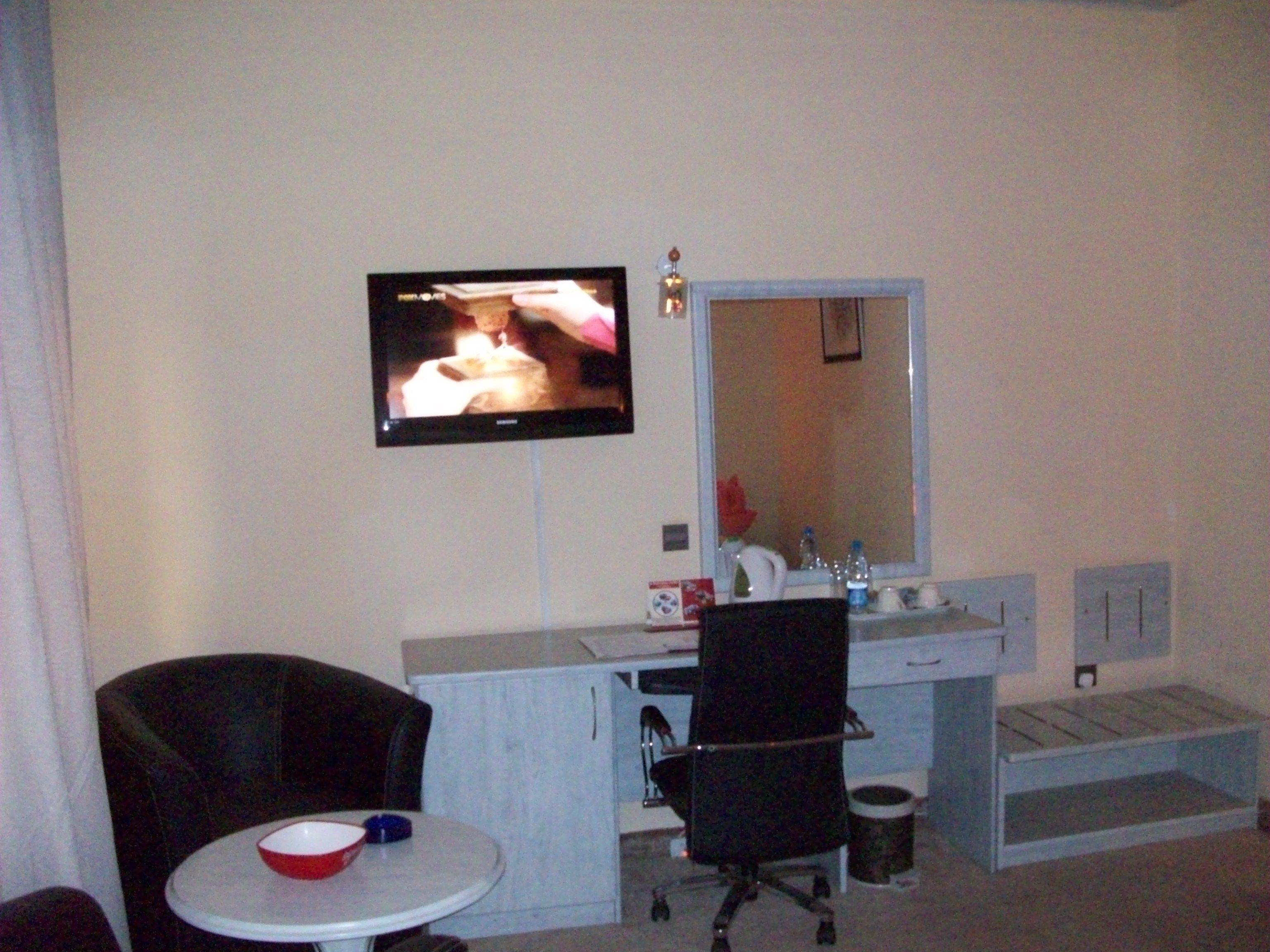 A hotel room with a television mounted on the wall, a desk with a mirror, a black office chair, a small round table with a red bowl, and a trash can.