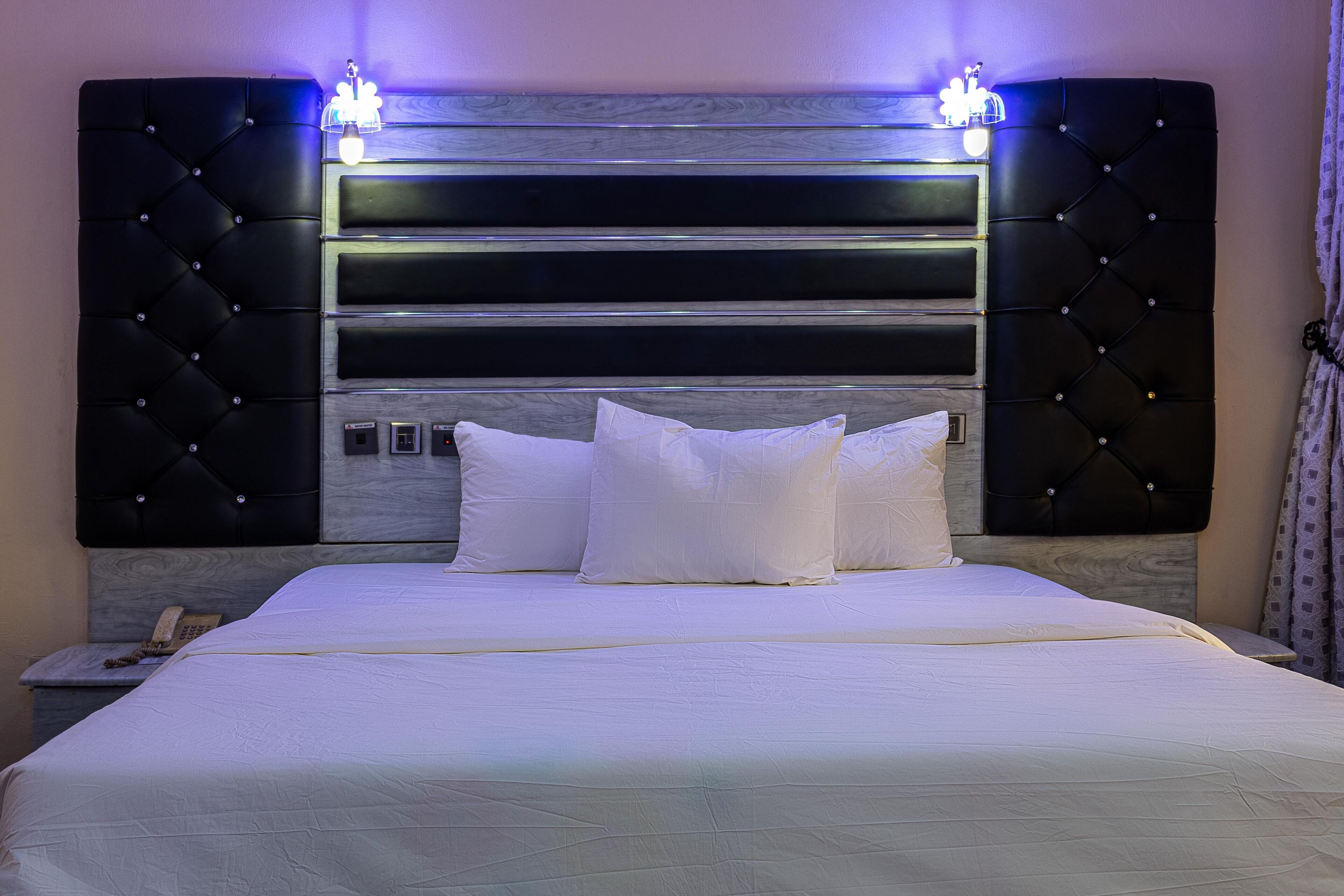 A neatly made bed with a tufted headboard, a bedside telephone, and a wall-mounted light fixture.
