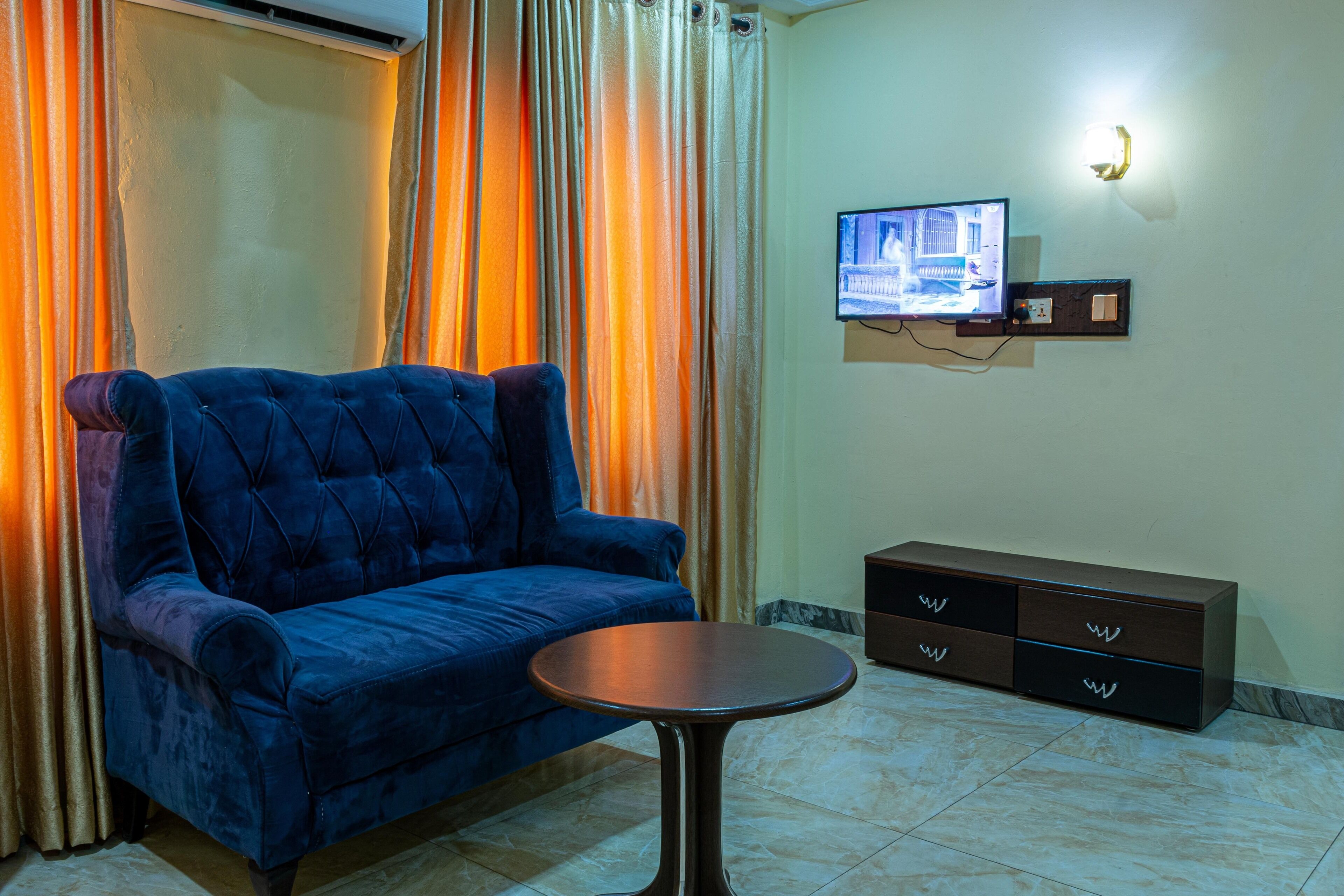 A room with a blue sofa, a small round table, a TV mounted on the wall, and a cabinet with drawers.