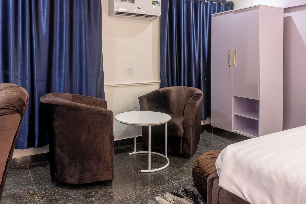 A hotel room with a bed, two brown armchairs, a small white table, a wardrobe, and blue curtains.
