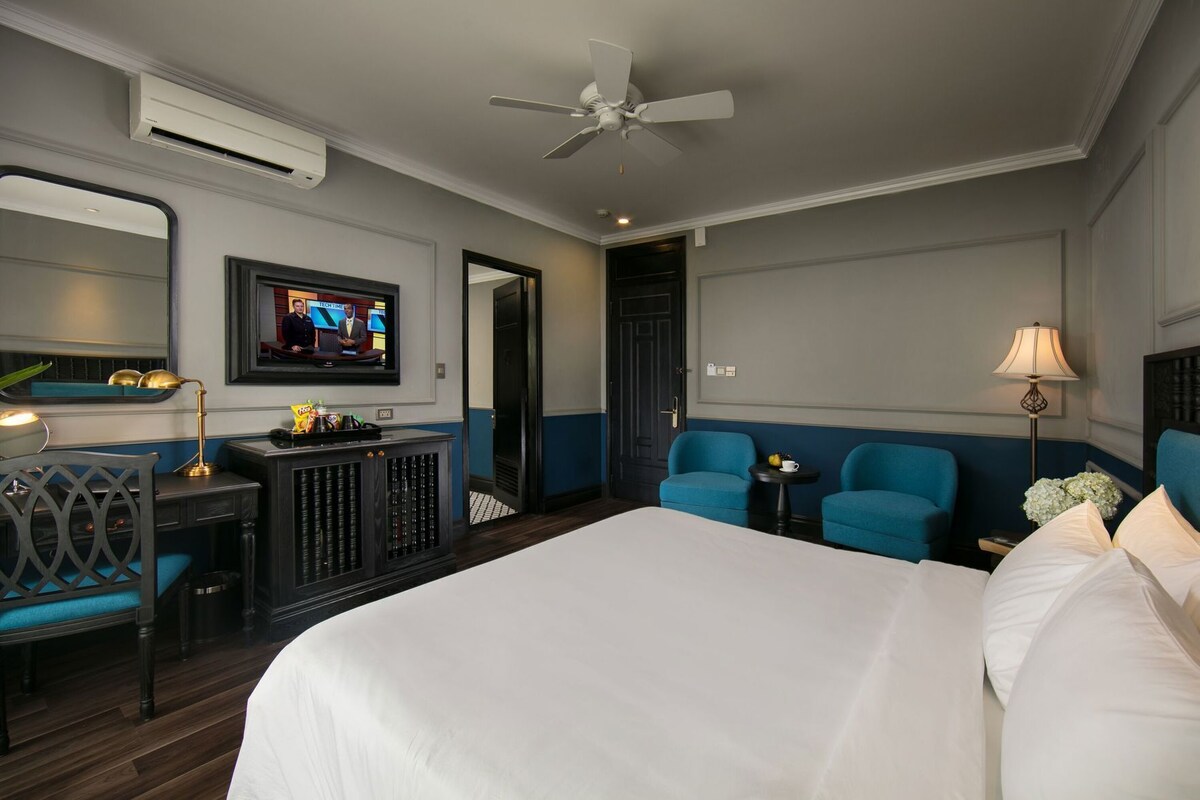 A hotel room with a bed, a desk with a computer, two chairs, a television, and a ceiling fan.