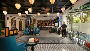 A hotel lobby with a modern design, featuring a reception desk, seating area with blue chairs, a patterned floor, and a large window offering a view of greenery. - Hoi An Golden Holiday Elite (Da Nang)