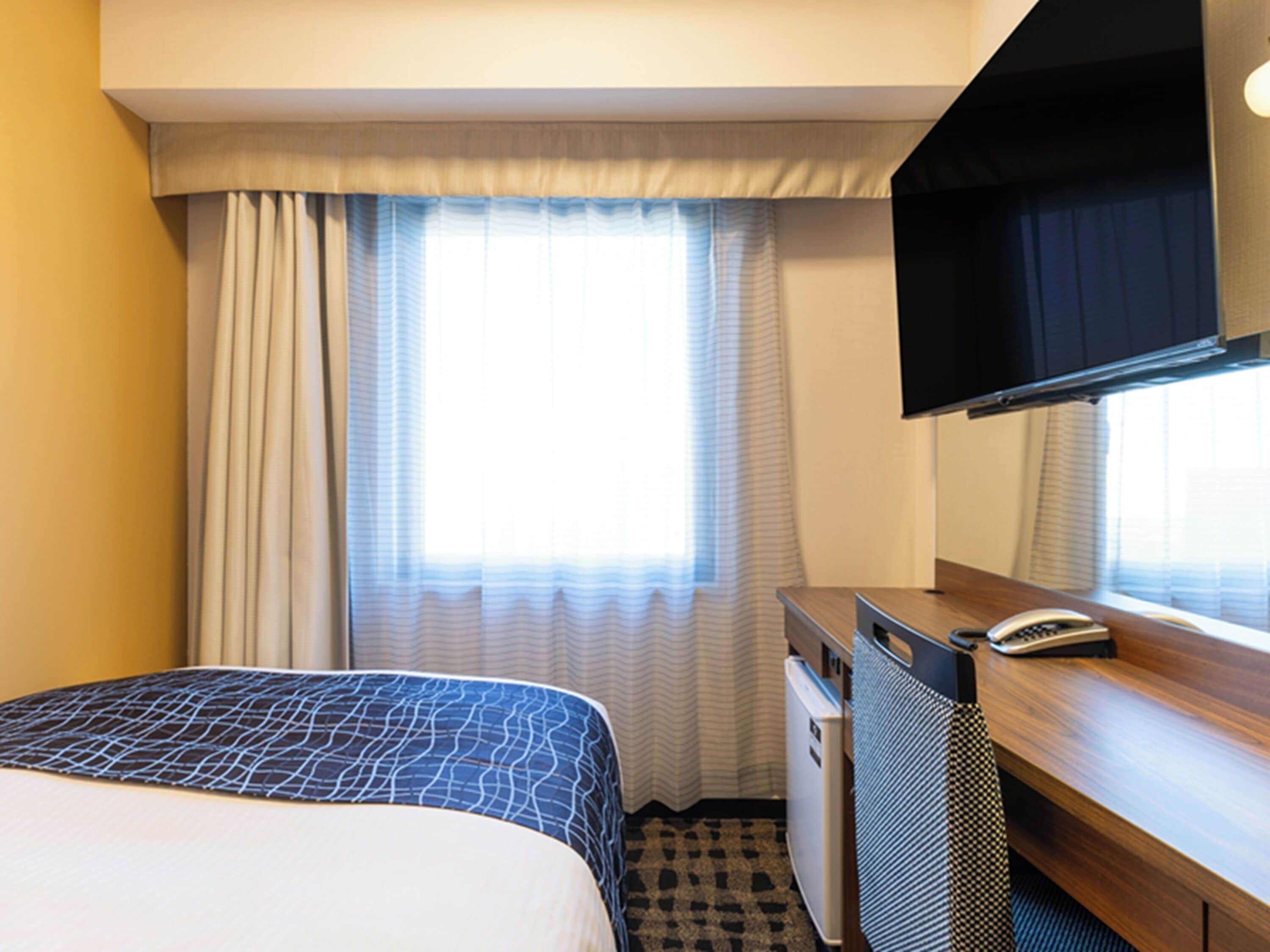 A hotel room with a bed, a desk with a telephone, a television, and a refrigerator.