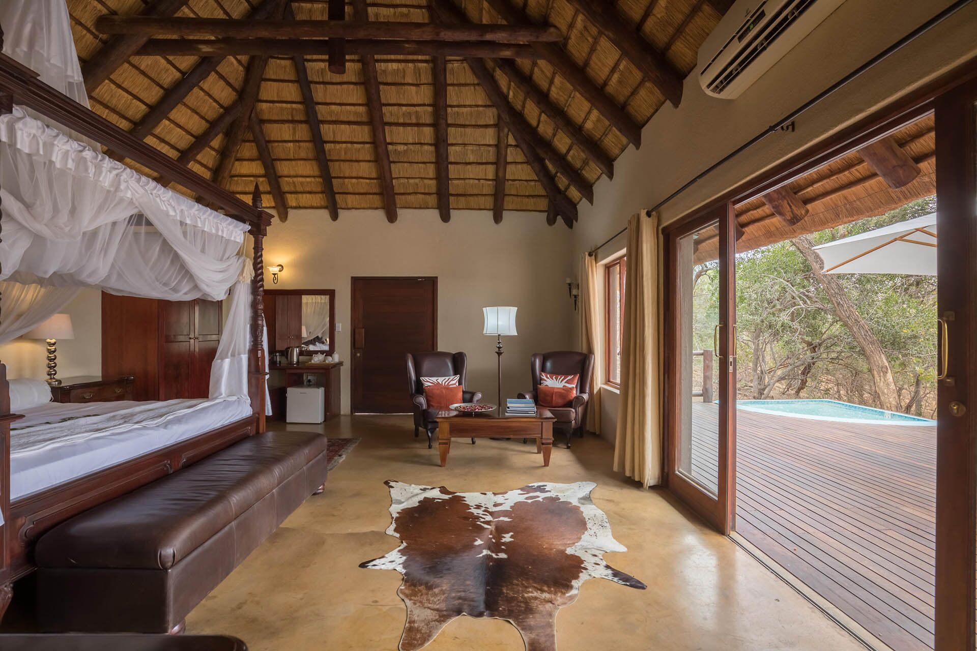 A room with a bed, a desk, chairs, a leather sofa, a cowhide rug, and a view of an outdoor pool.