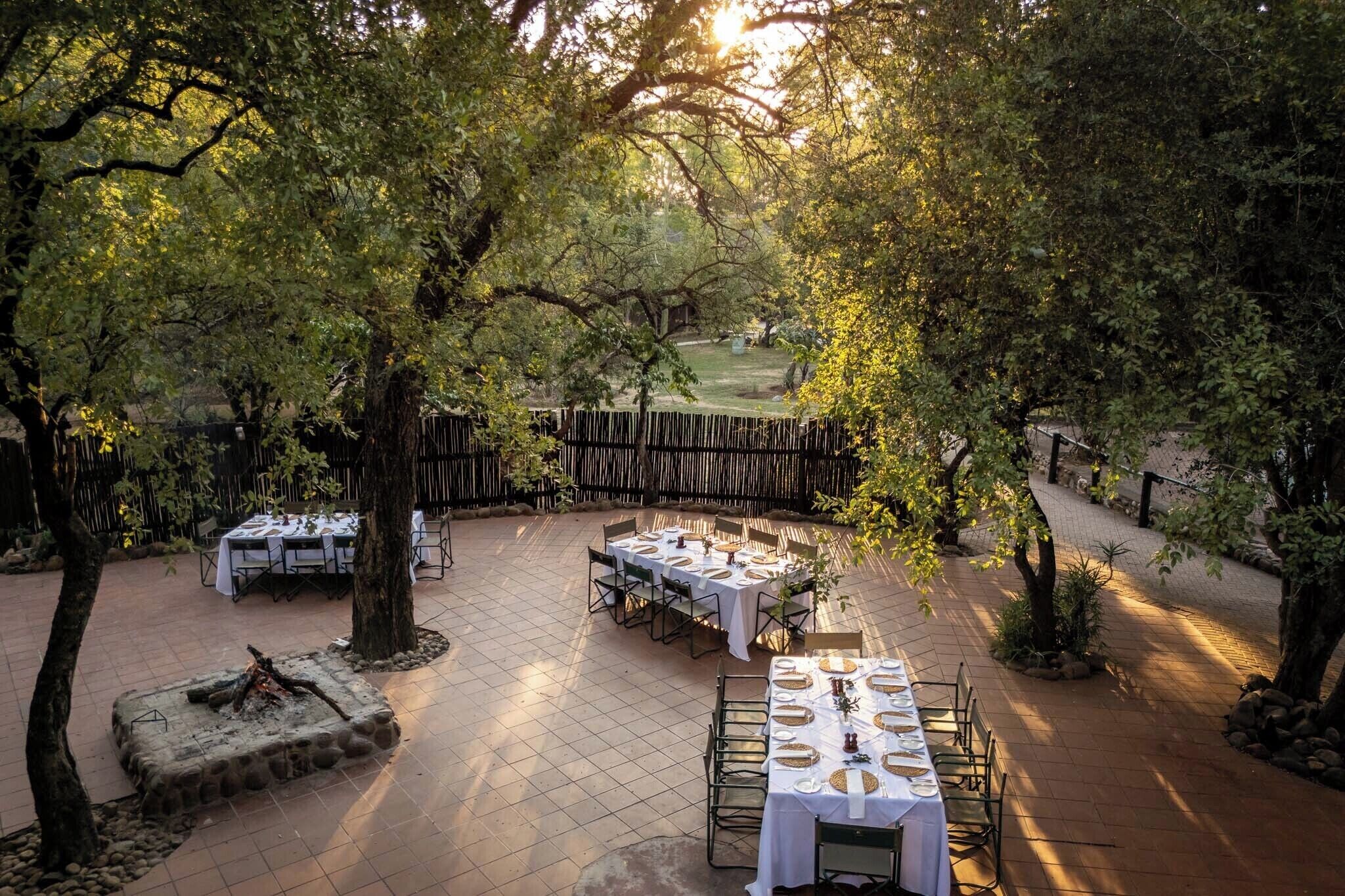 An outdoor dining area with multiple tables set up for a meal, surrounded by trees and a wooden fence.
