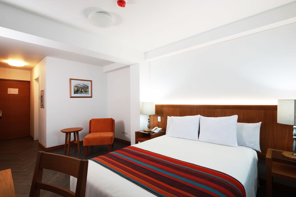 A hotel room with a bed, a chair, a small table, a nightstand with a lamp, and a wall-mounted telephone.