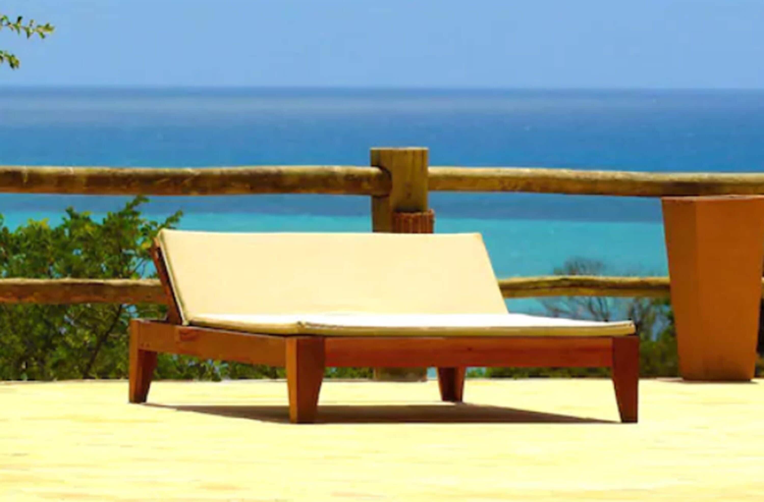 A wooden lounge chair with a cushion, positioned on a wooden deck with a view of the ocean.