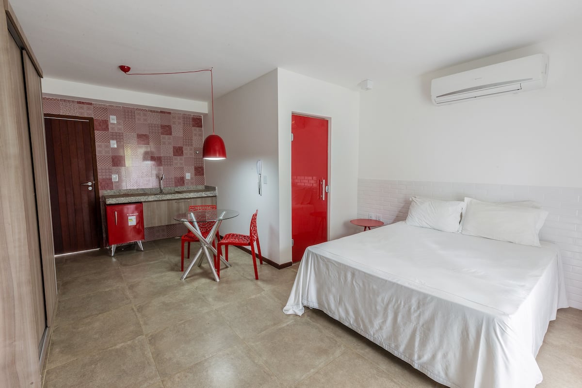 A hotel room with a bed, a small table with a chair, a red door, and a kitchen area with a red chair.
