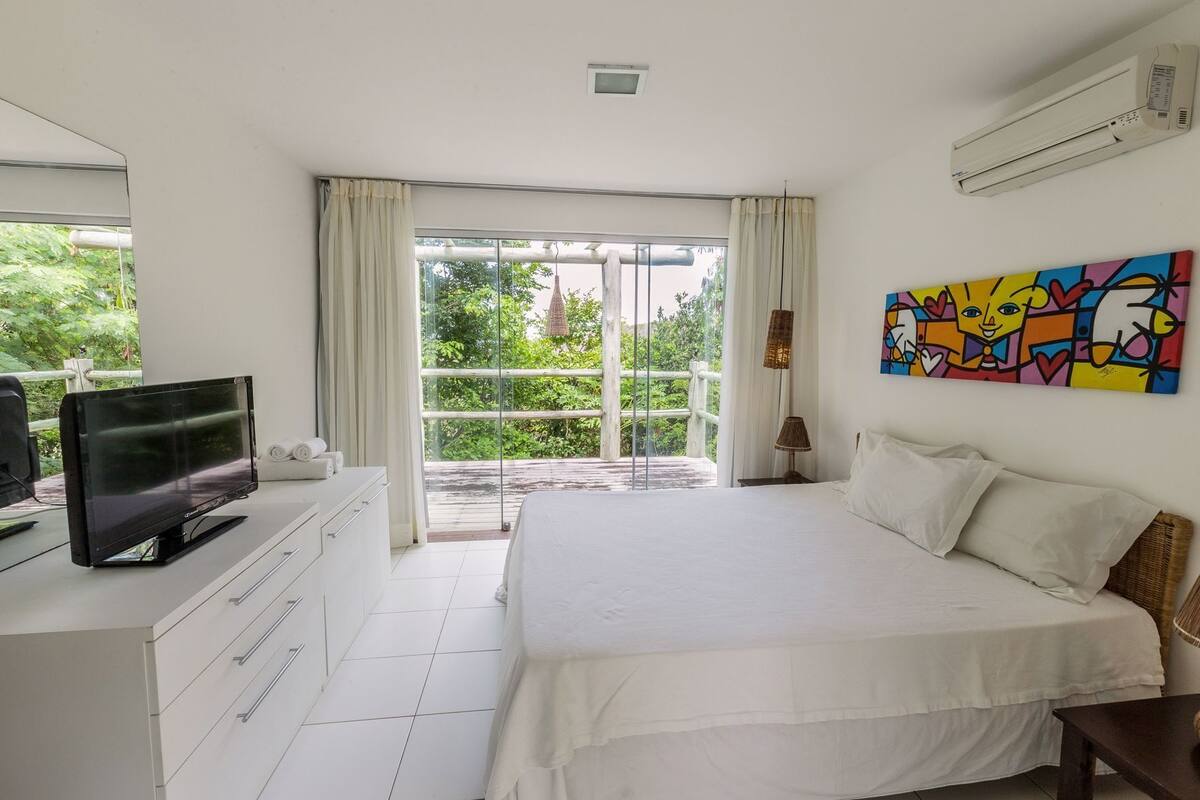 A bedroom with a bed, a TV, a painting, and a balcony with a view of greenery.