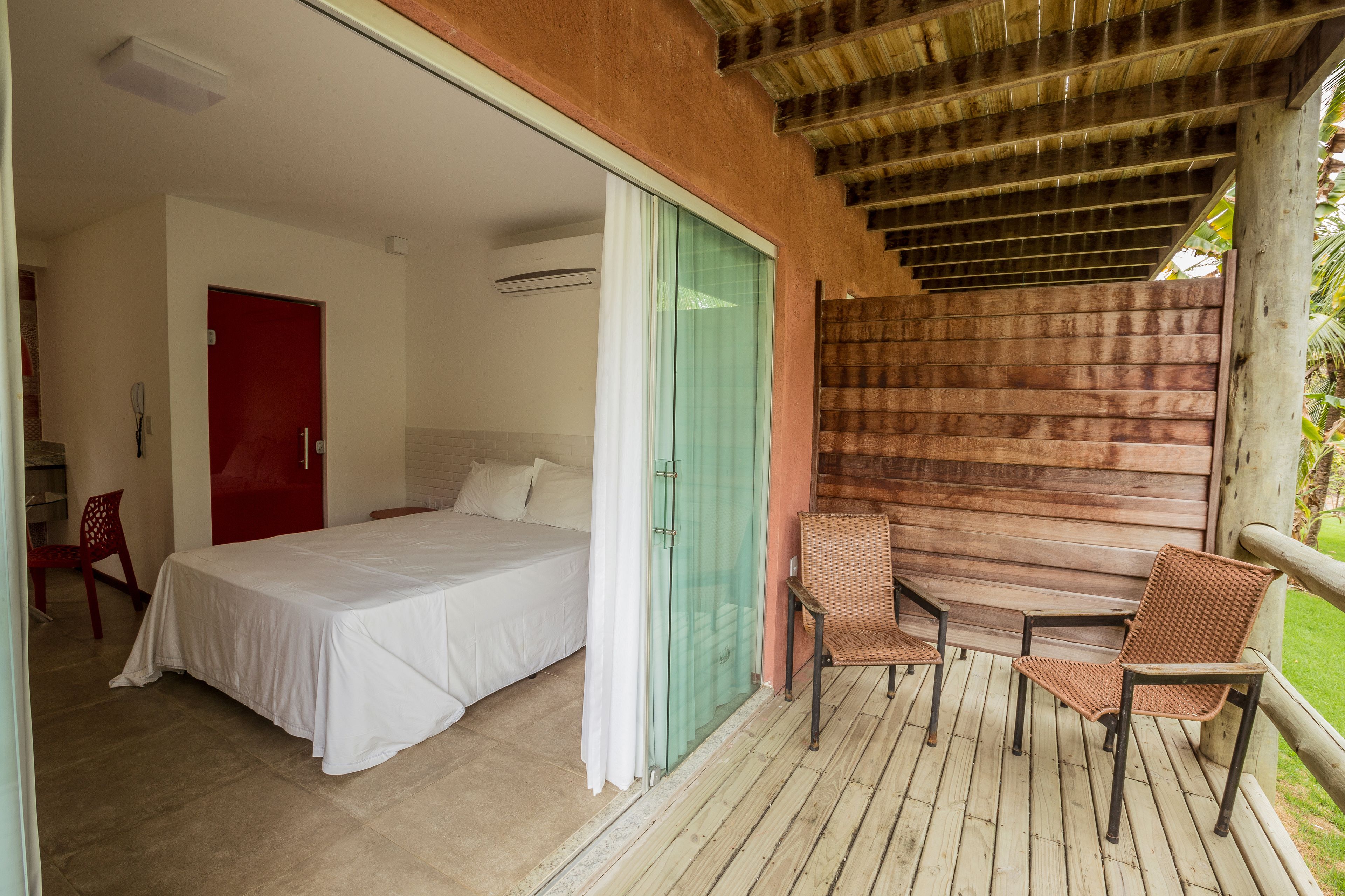 Ein Schlafzimmer mit einem Bett, einer roten Tür und einer Glastür, die auf einen Balkon mit Korbstühlen und einem Holztisch führt.