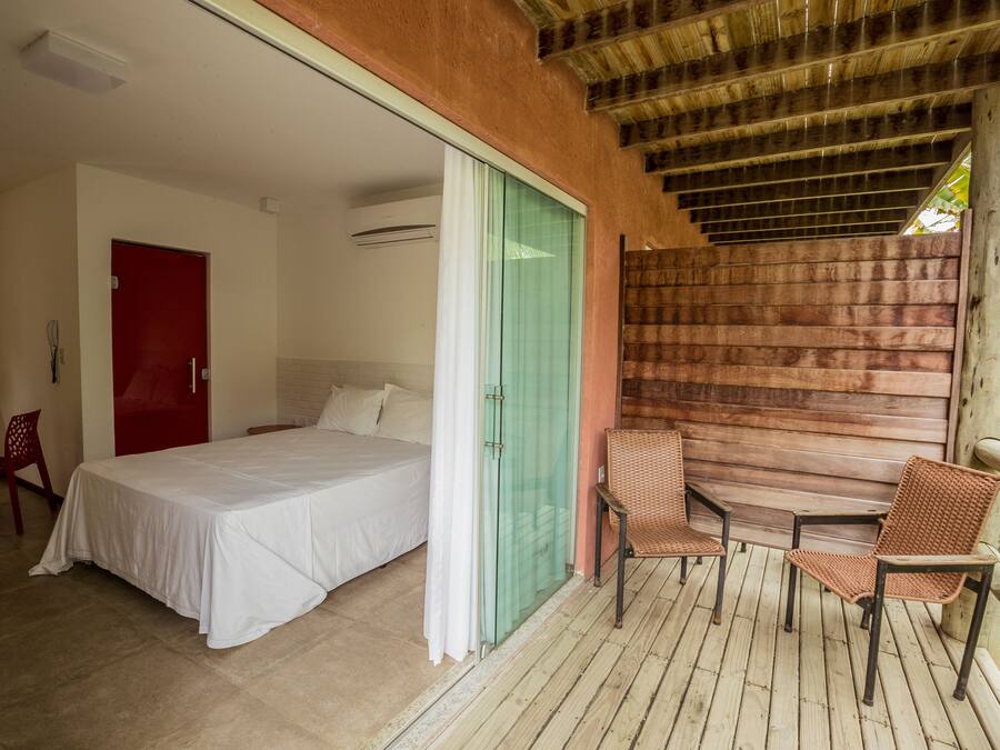 A bedroom with a bed, a red door, and a sliding glass door leading to a balcony with wicker chairs and a wooden table.