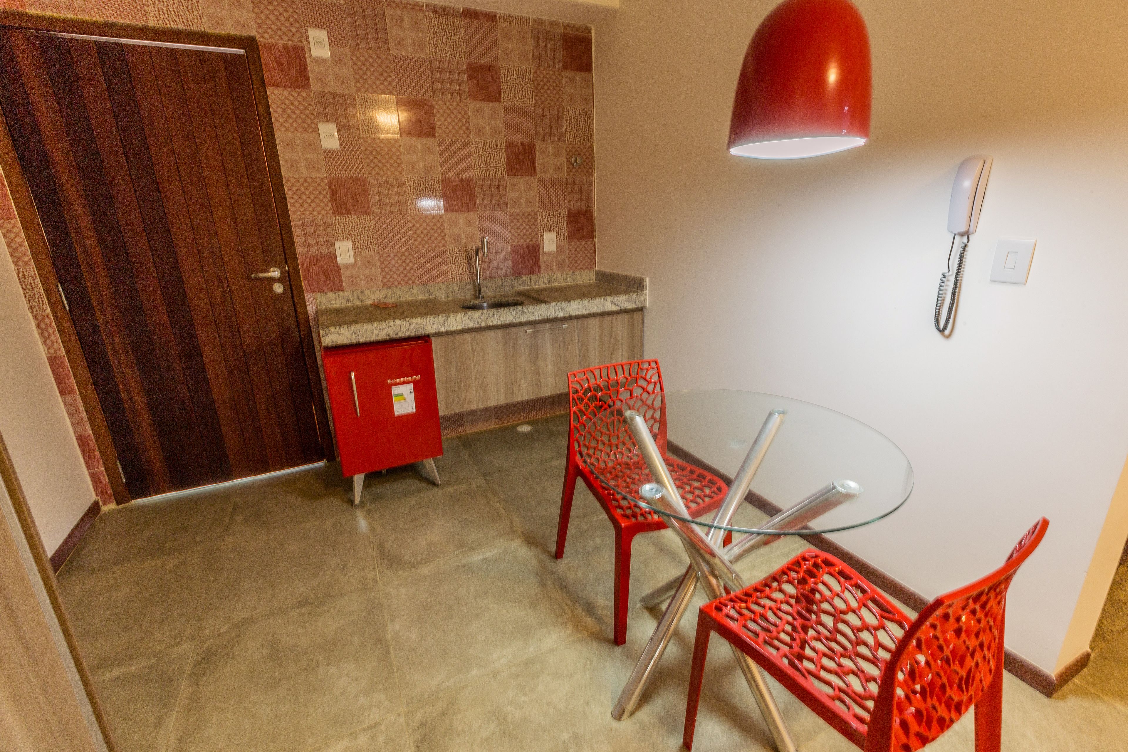 Una habitación con puerta de madera, salpicadero de azulejos, un armario rojo, una mesa de vidrio y sillas rojas.