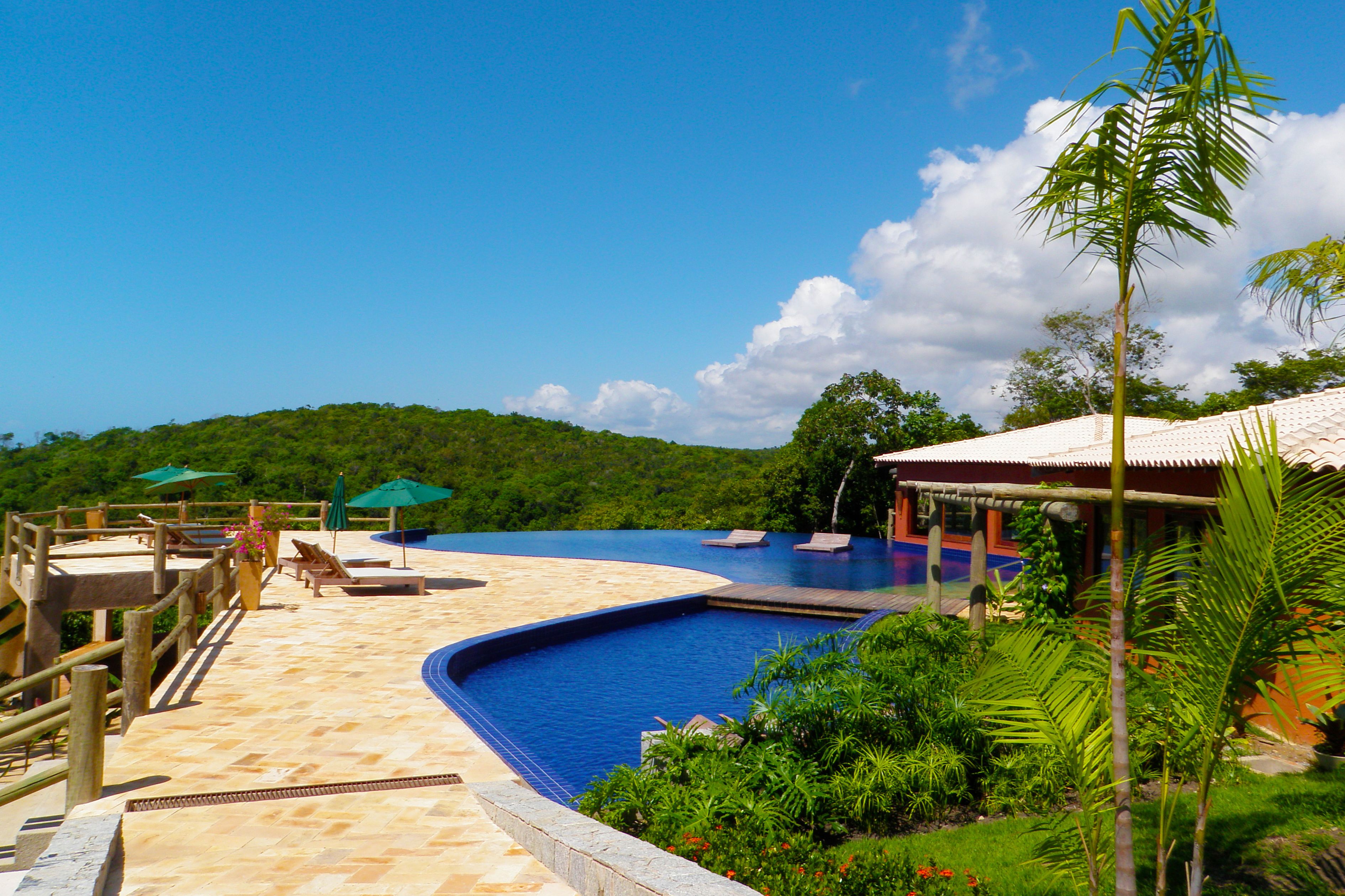 Una piscina con sillas de descanso y sombrillas, rodeada de abundante vegetación y una terraza de madera.