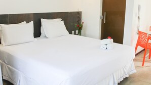 Una habitación de hotel con una cama grande, ropa de cama blanca, un cabecero de madera, una mesita con un jarrón de flores y una silla roja.