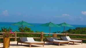Una terraza con tres sillones de descanso y dos sombrillas verdes, con vistas al mar azul y despejado.