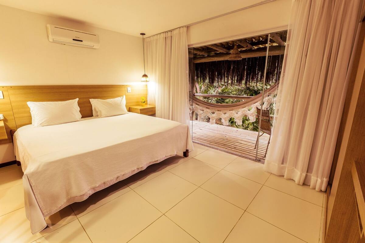 A hotel room with a bed, bedside table, lamp, air conditioning unit, and a view of a hammock and greenery outside.