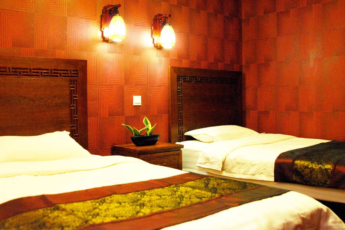 A hotel room with two beds, a nightstand with a plant, and wall-mounted lights.