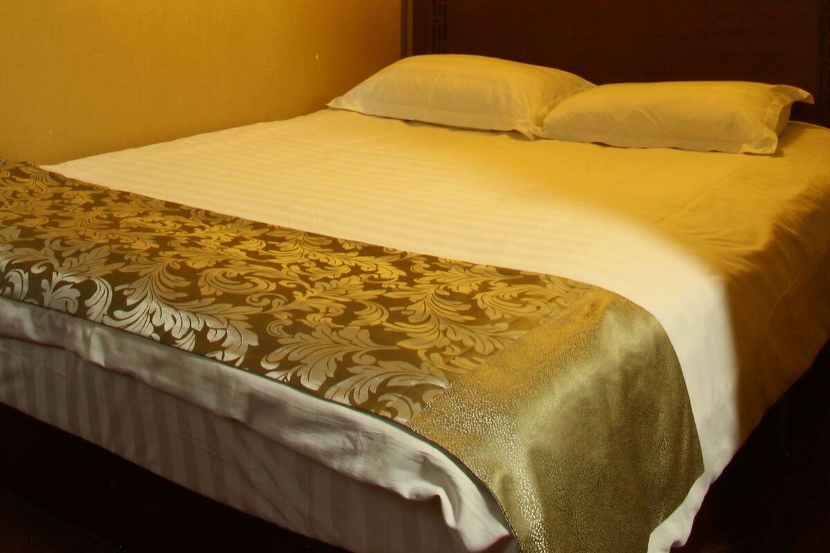 A hotel room with a bed, a headboard with a decorative pattern, and a wall-mounted light fixture.