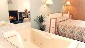 Habitación de hotel con una bañera grande, una cama con cubrecama estampado, una mesita de noche con lámpara y un espejo que refleja una chimenea.