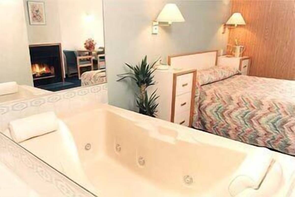Habitación de hotel con una bañera grande, una cama con cubrecama estampado, una mesita de noche con lámpara y un espejo que refleja una chimenea.