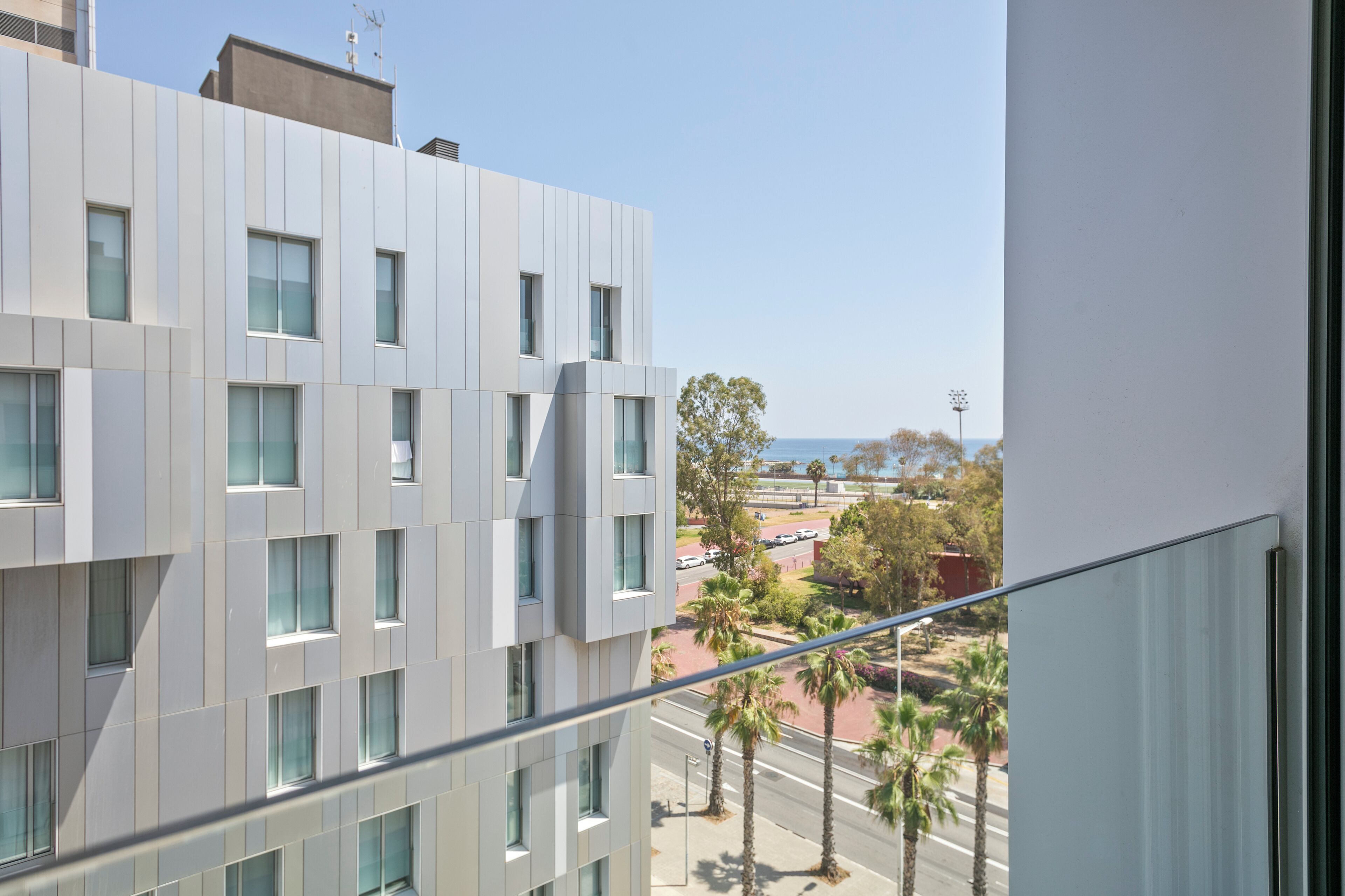 A modern building with a balcony overlooking a street with palm trees and parked cars.