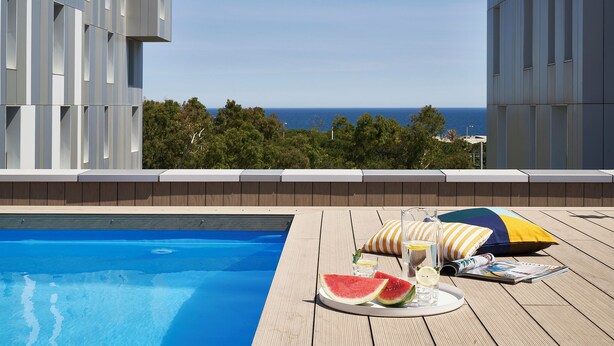 Zona de piscina con vista al mar, una terraza de madera y una mesa con sandía, vasos y una revista.