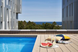 Zona de piscina con vista al mar, una terraza de madera y una mesa con sandía, vasos y una revista.
