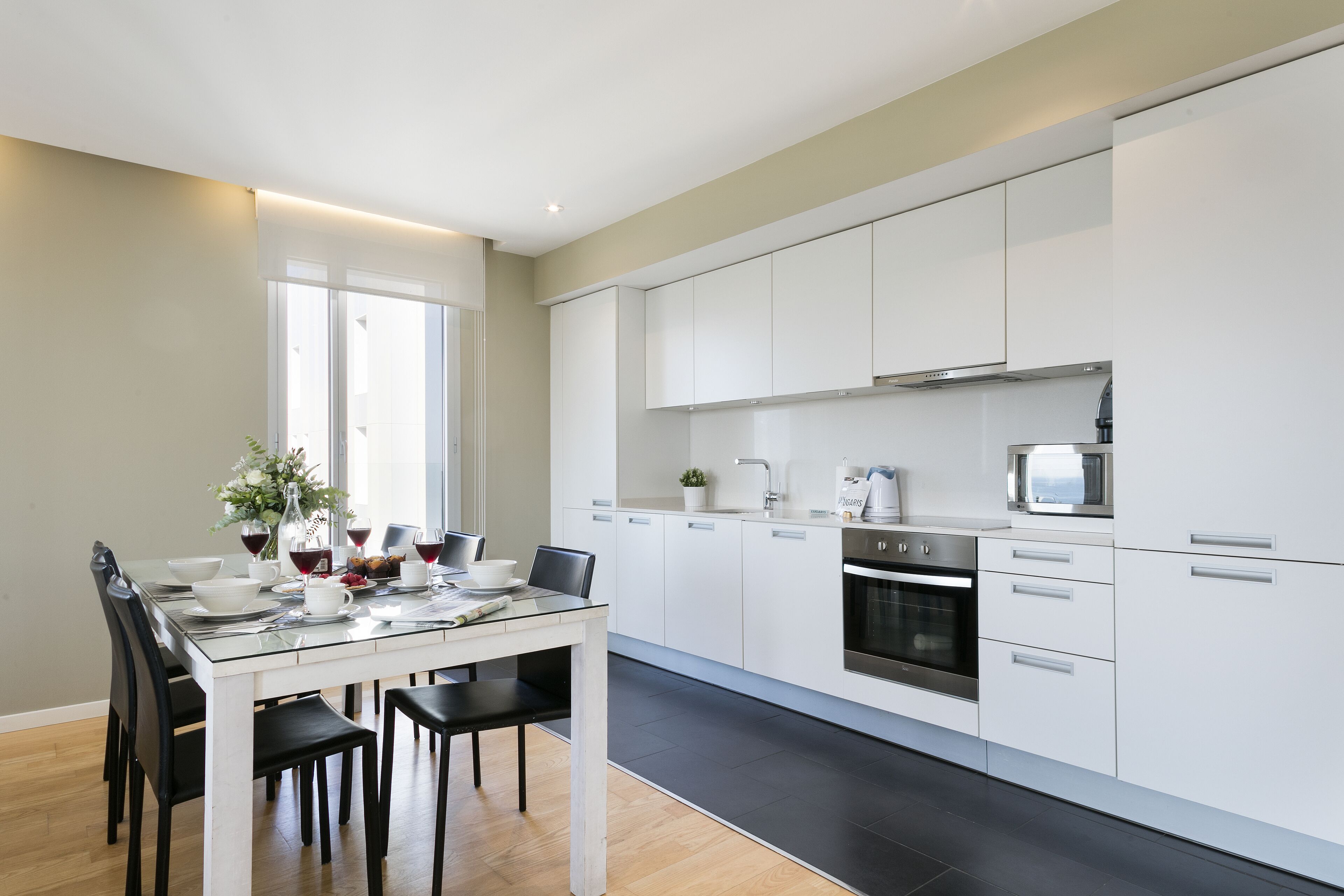 A modern kitchen with white cabinets, a dining table with chairs, and a large window providing natural light.