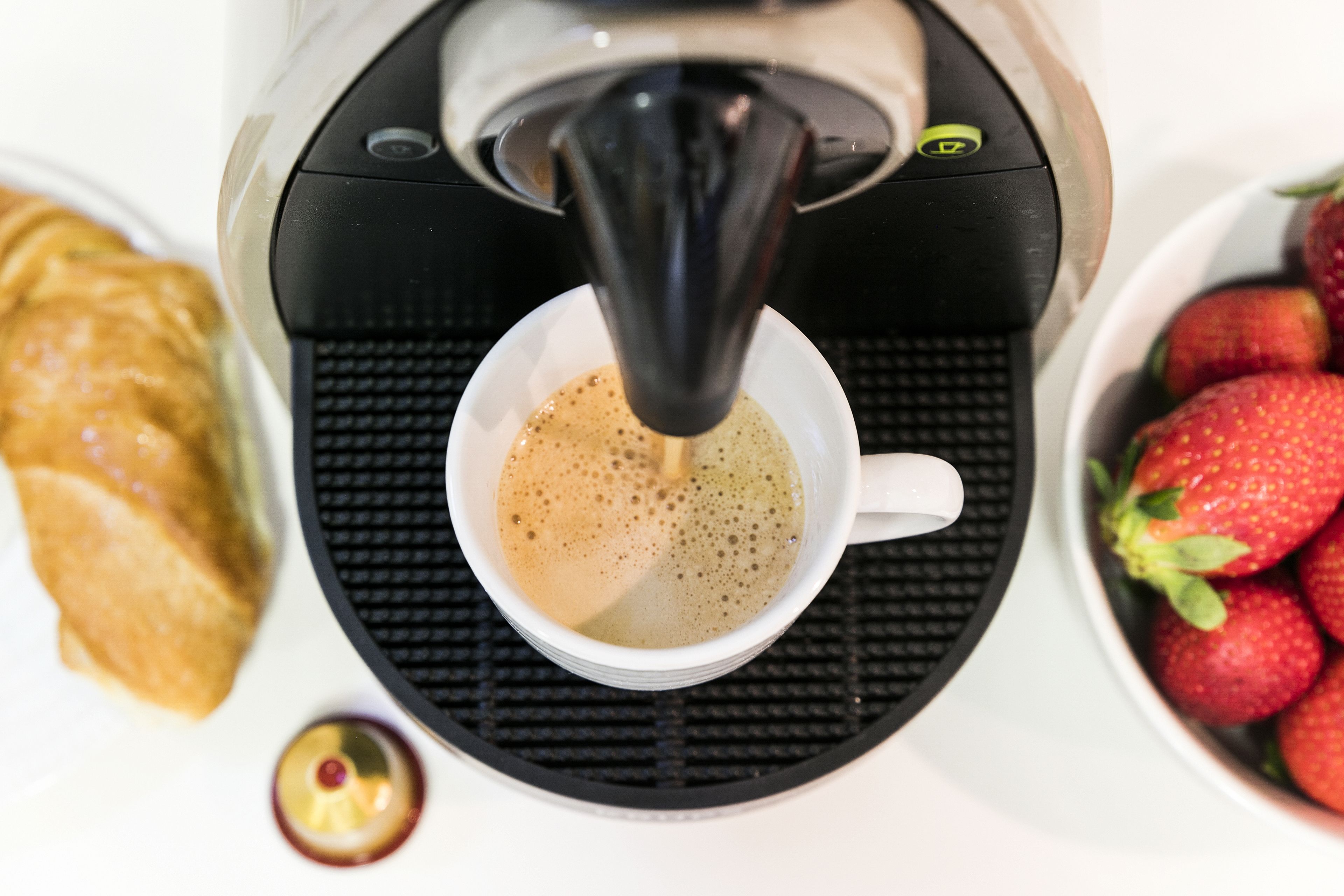 A coffee machine dispensing coffee into a cup, a croissant, and a bowl of strawberries.
