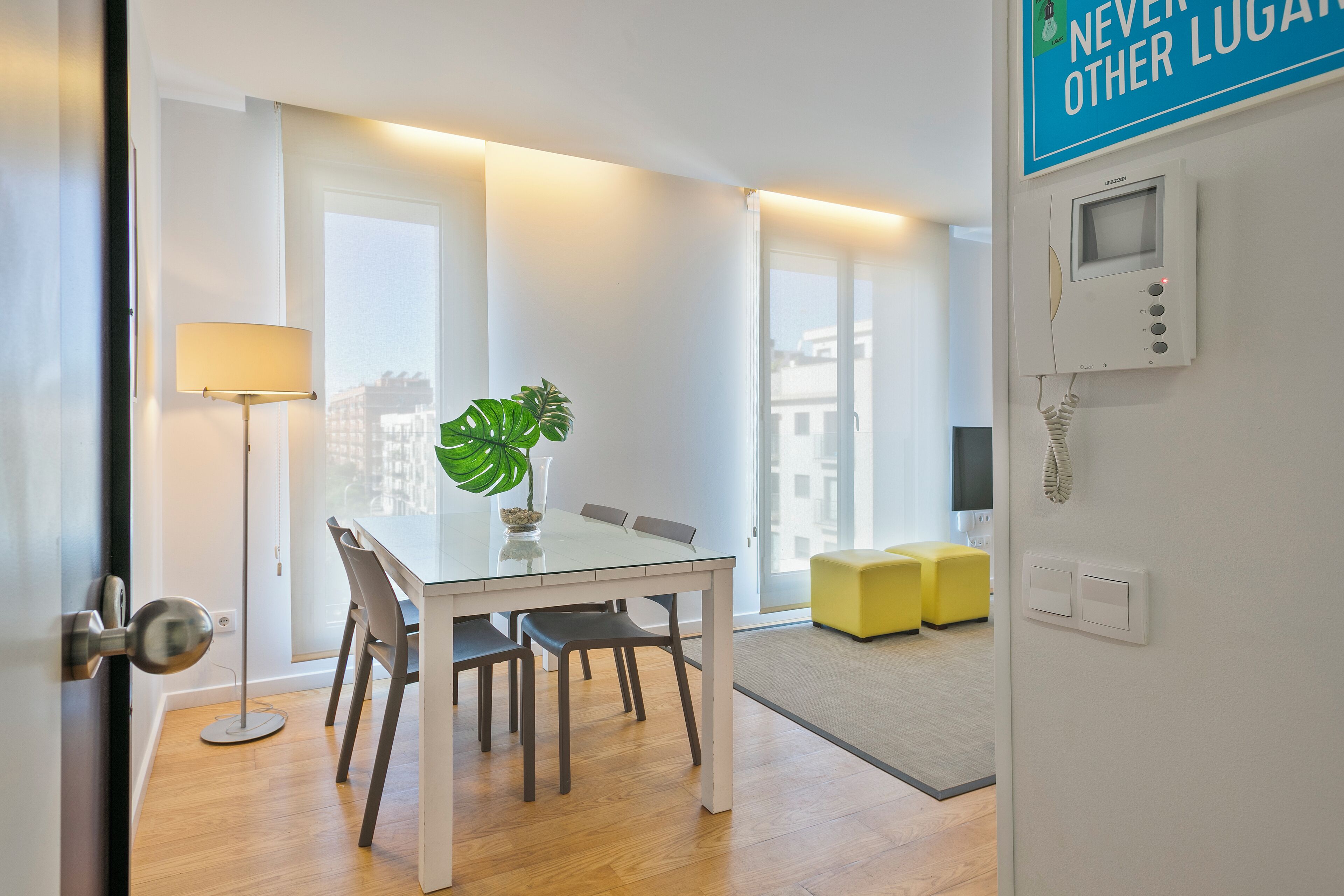 A modern dining area with a glass table, chairs, a potted plant, and a yellow ottoman.
