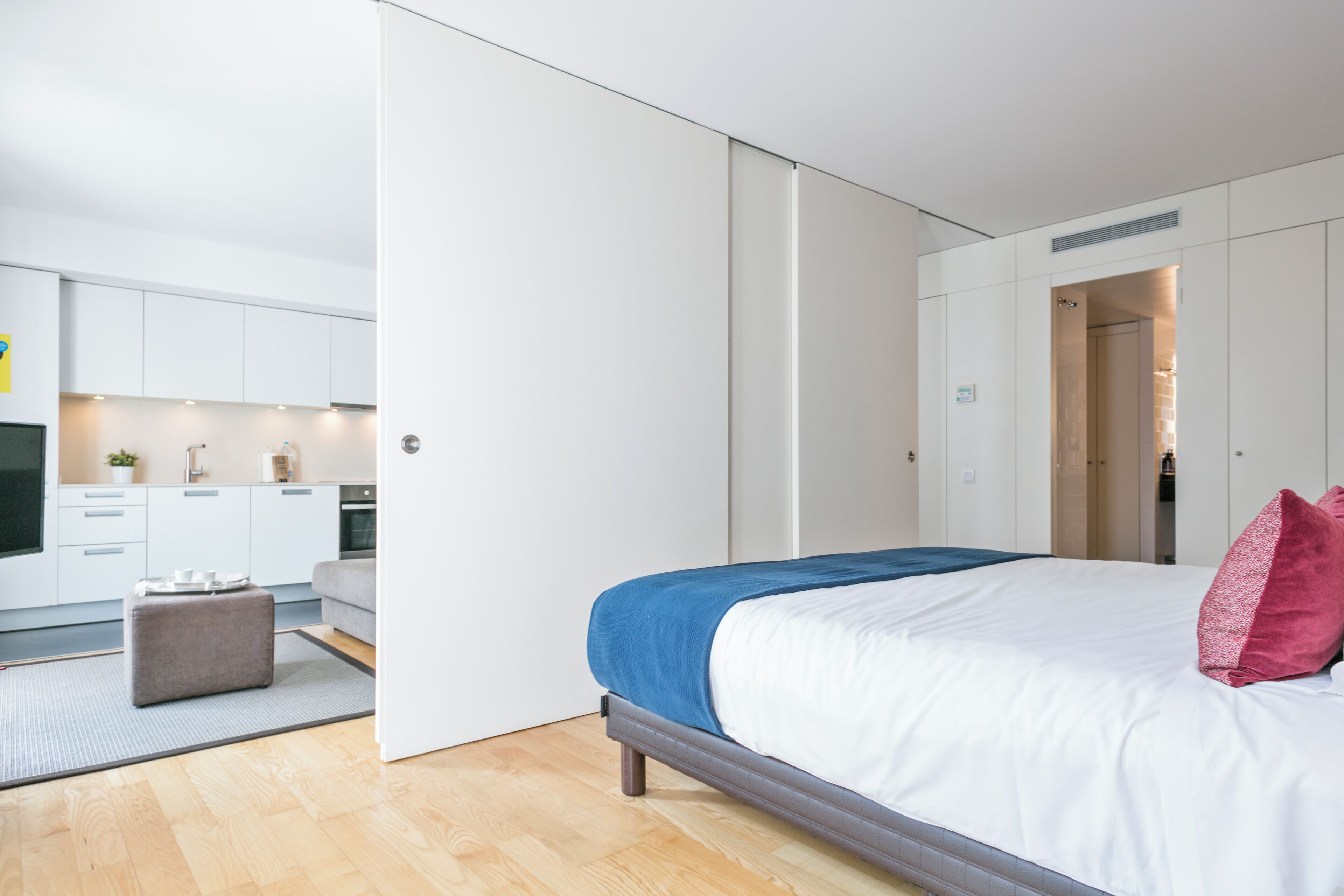 A modern bedroom with a large bed, a small seating area, and a visible kitchen area.