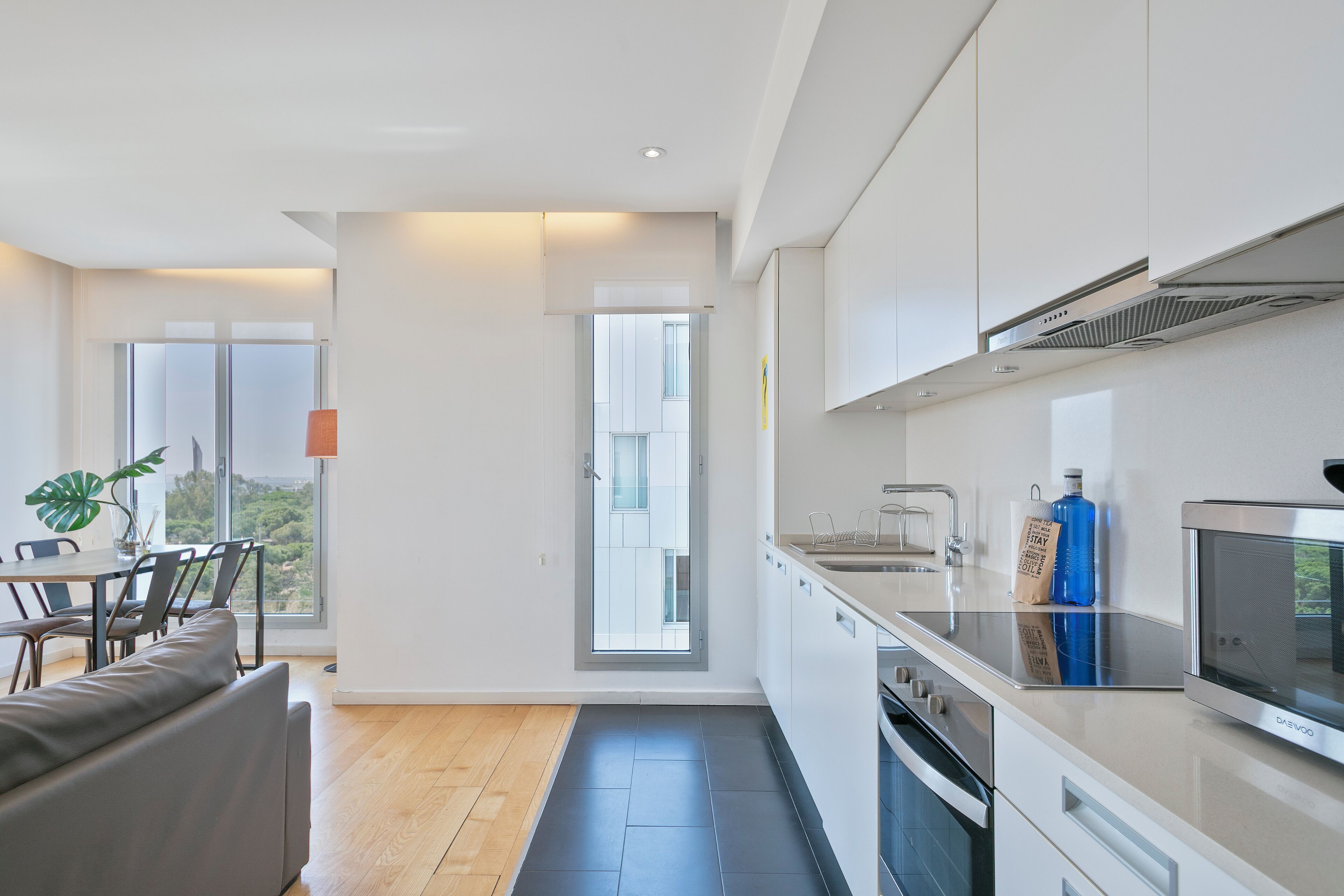 A modern kitchen with white cabinets, a built-in oven, and a microwave. There is a dining area with a table and chairs, and a large window providing a view of the outdoors.