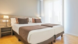 Una habitación de hotel moderna con dos camas, una mesita de noche y un ventanal con cortinas traslúcidas.