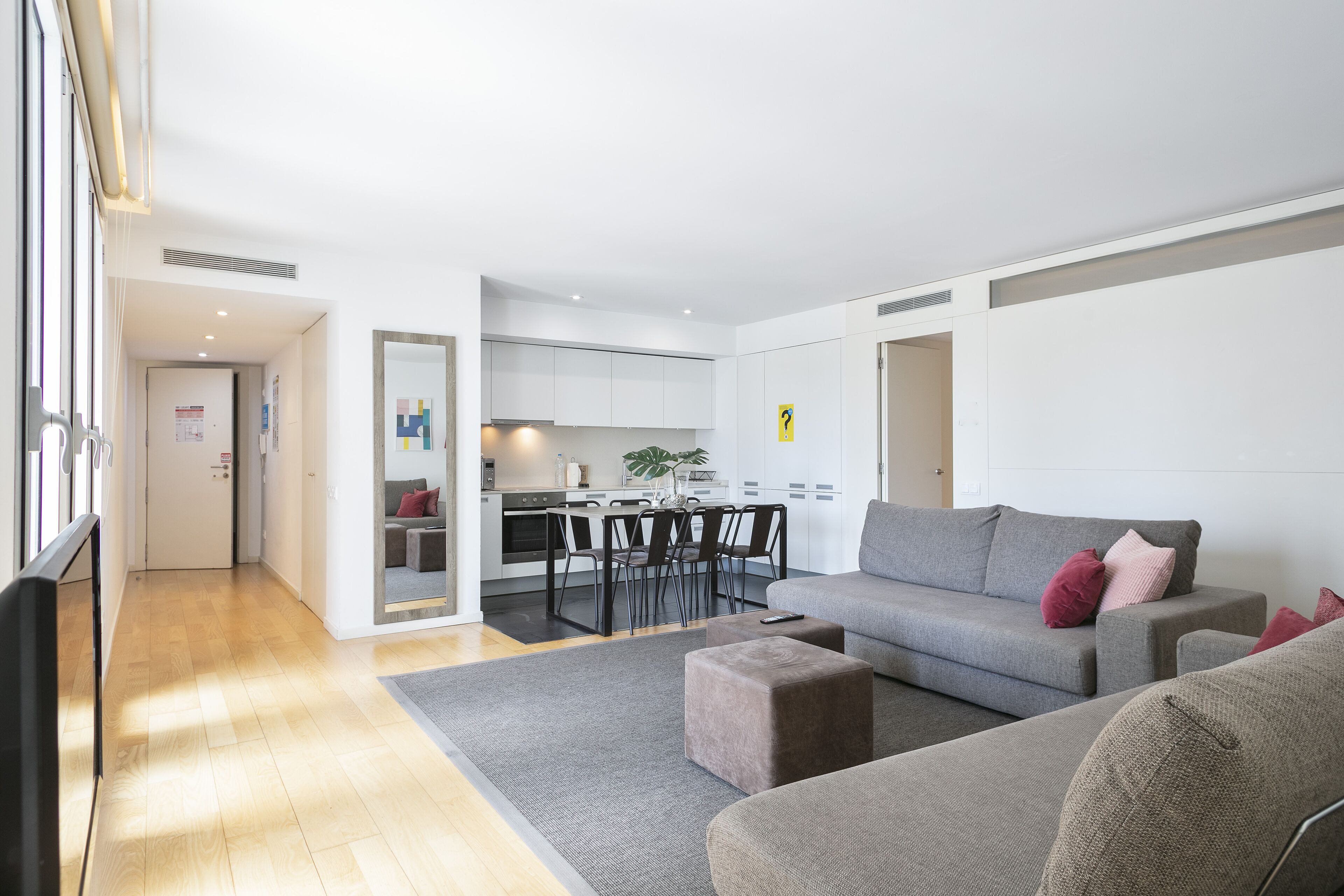 Una sala moderna con un sofá gris, un otoman y un comedor, con una cocina al fondo.