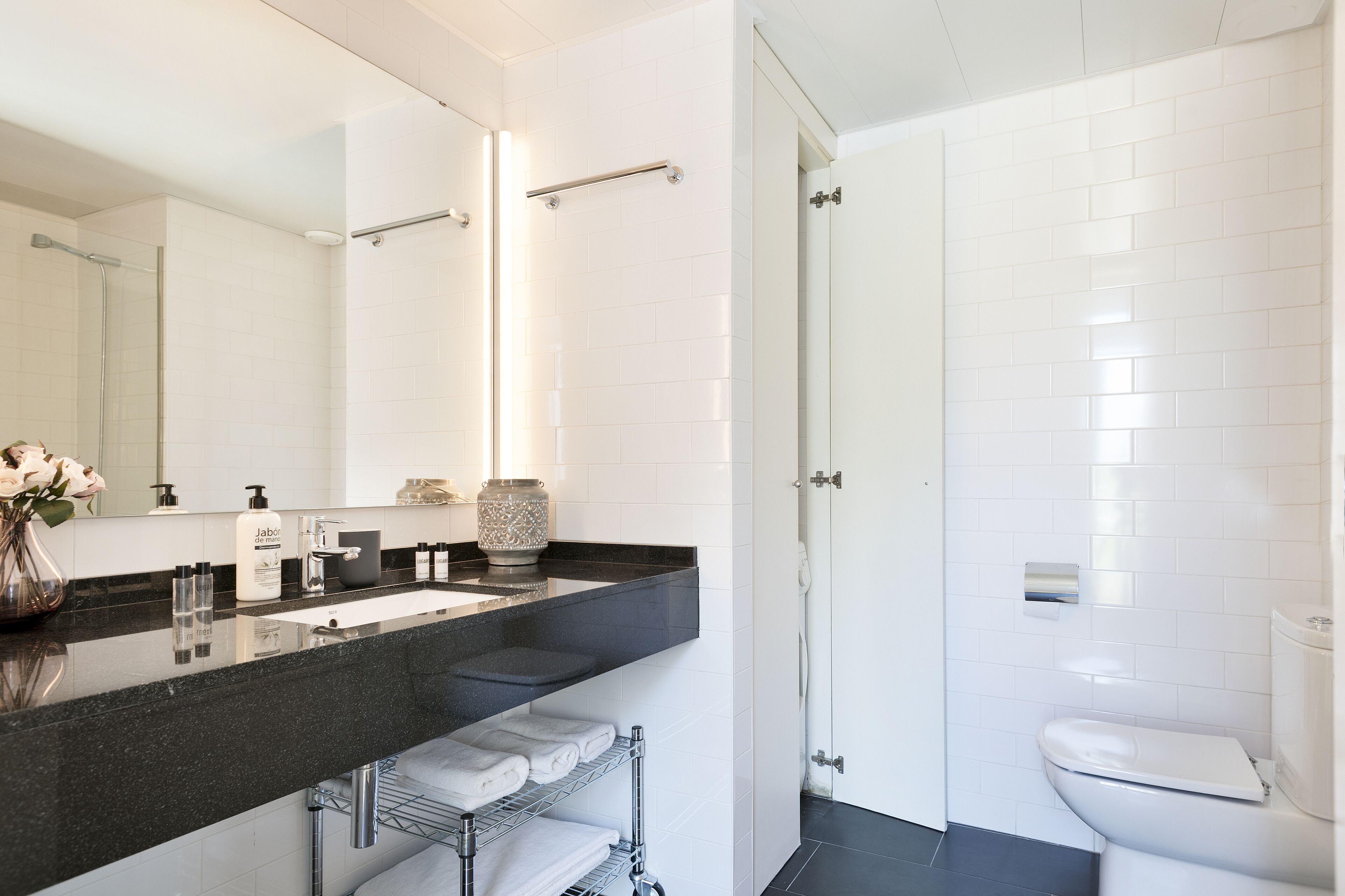 A modern bathroom with a black countertop, white tiles, a glass shower enclosure, and a white toilet.