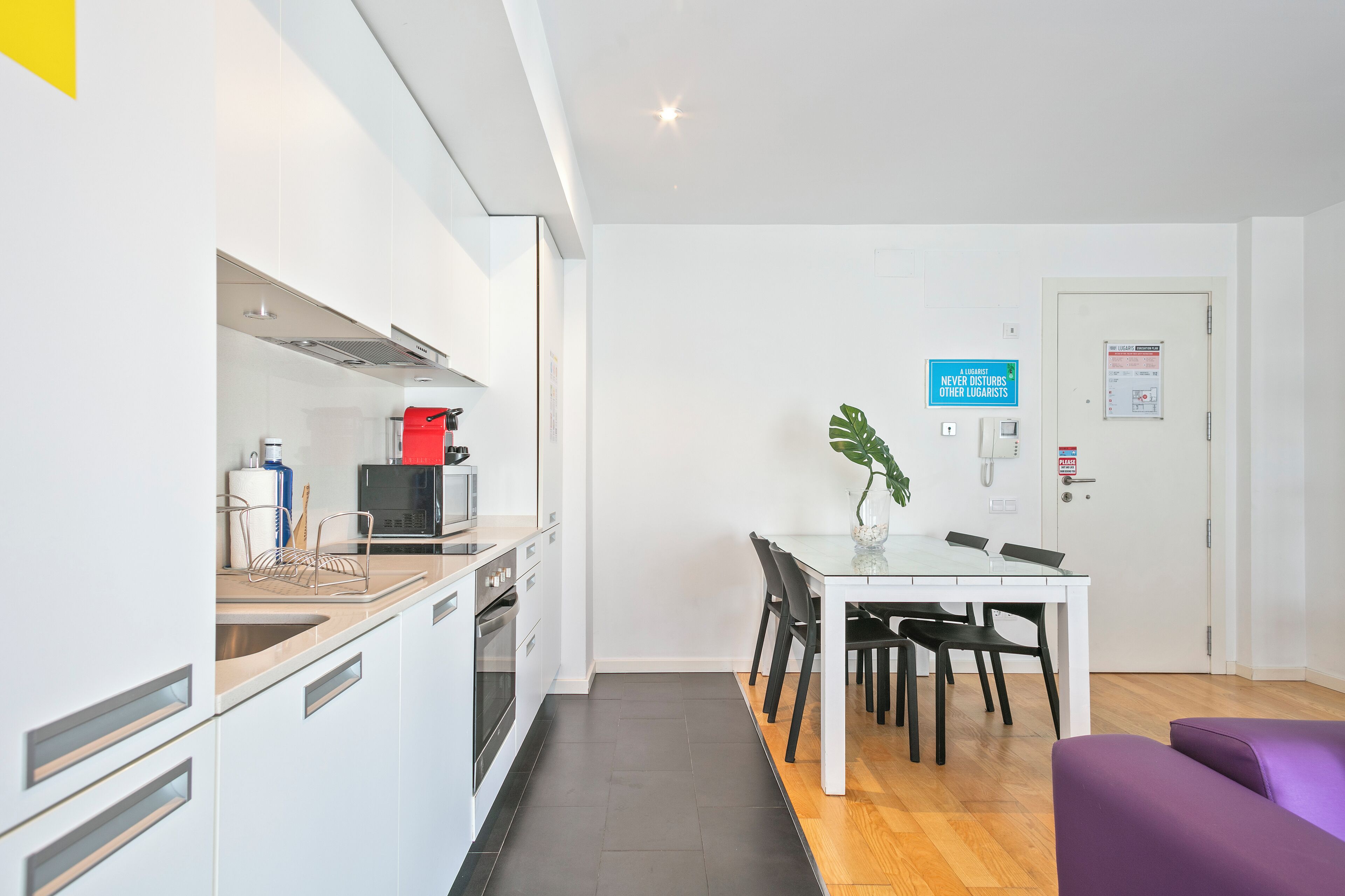 A modern kitchen with white cabinets, a microwave, and a red kettle. There is a dining table with black chairs and a purple sofa.