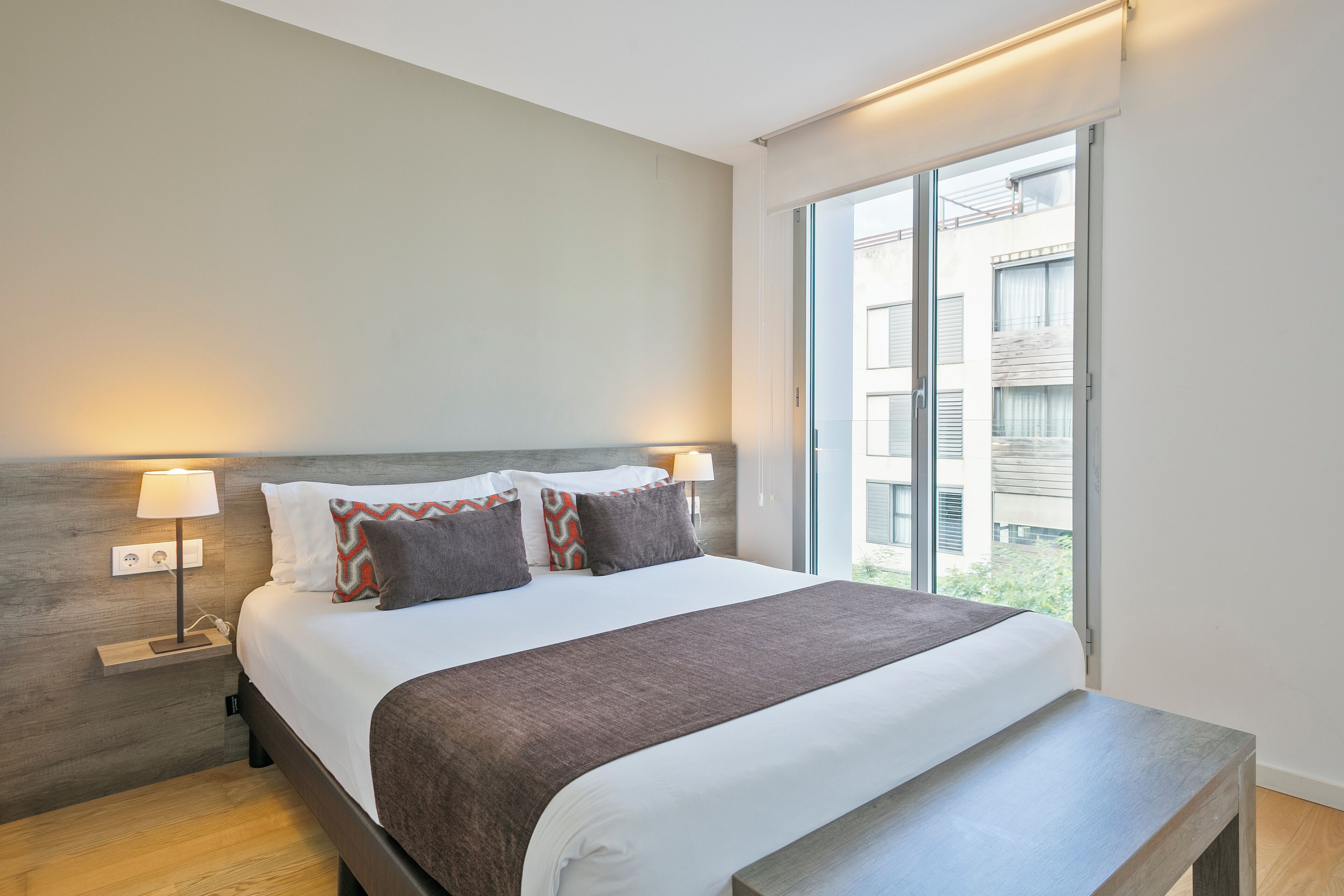 Een moderne hotelkamer met een groot bed, nachtlampjes, een bankje en uitzicht op de stad door grote ramen.