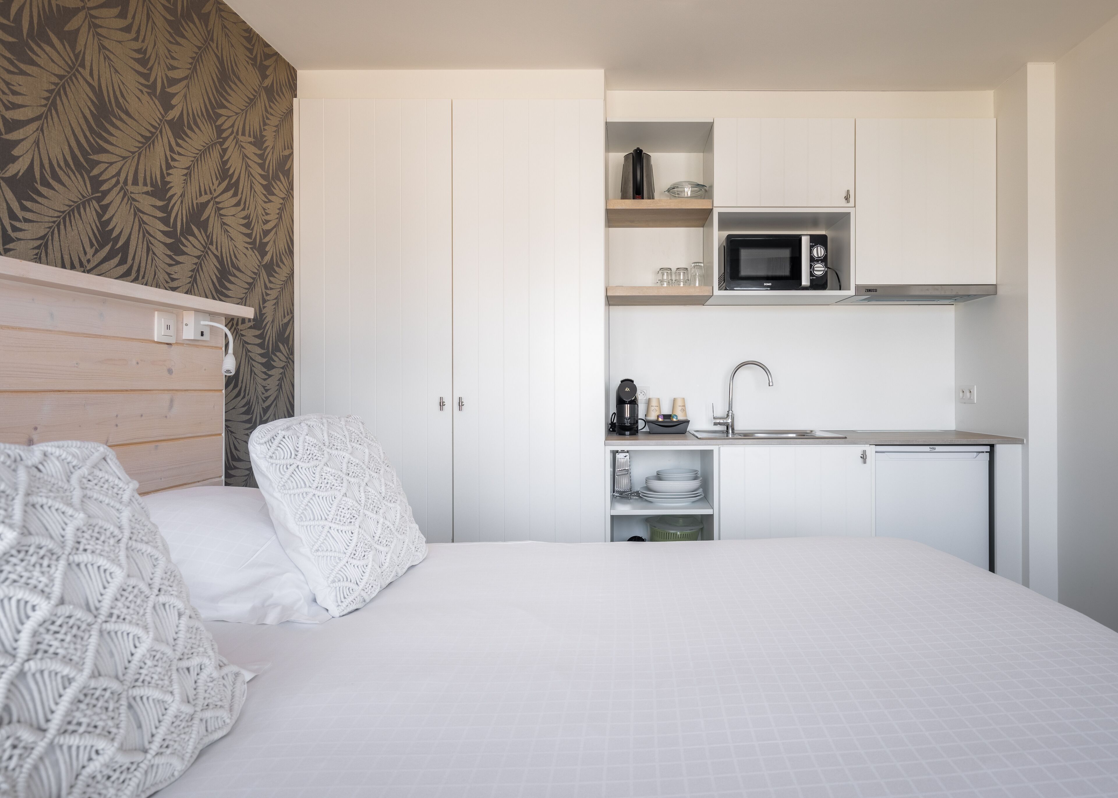 A compact bedroom with a bed, a bedside table, and a small kitchenette with a microwave and sink.
