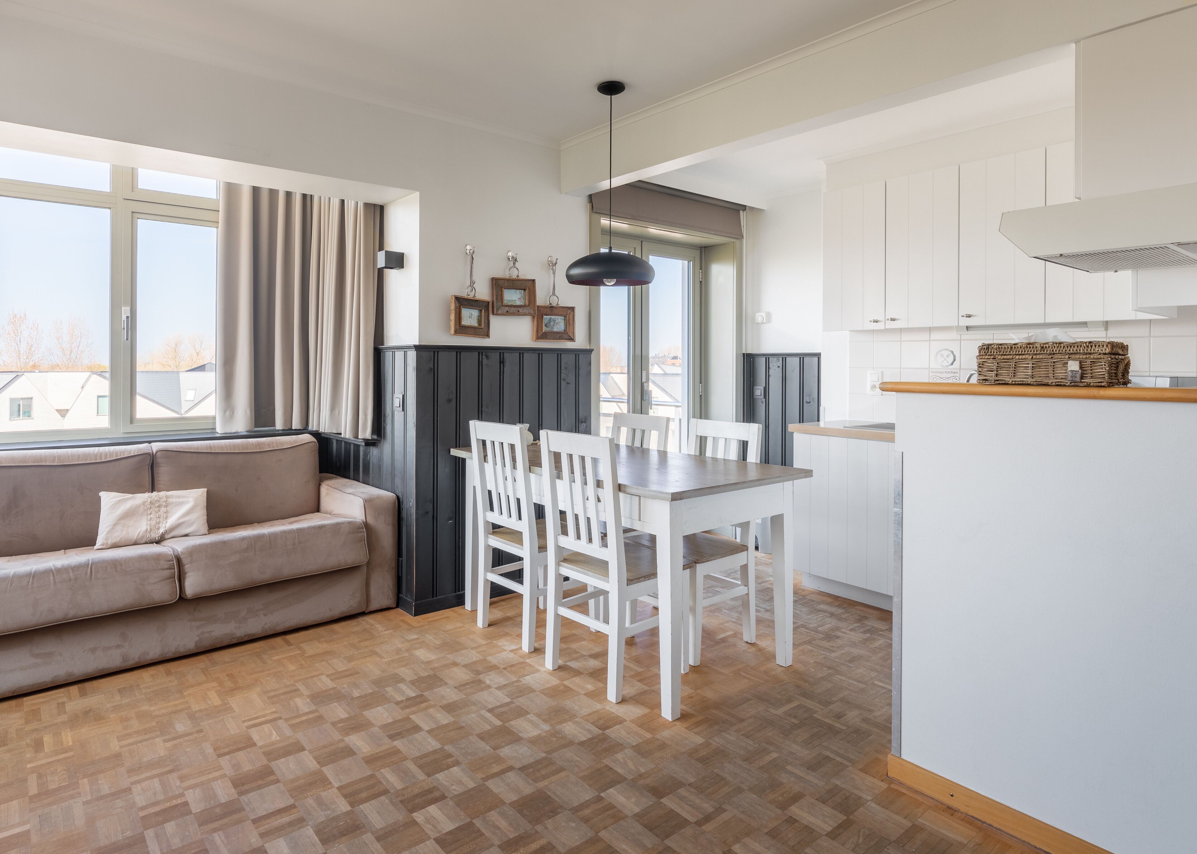 Une cuisine moderne avec des meubles blancs, un coin repas avec des chaises blanches et un parquet en bois disposé en motif chevron.