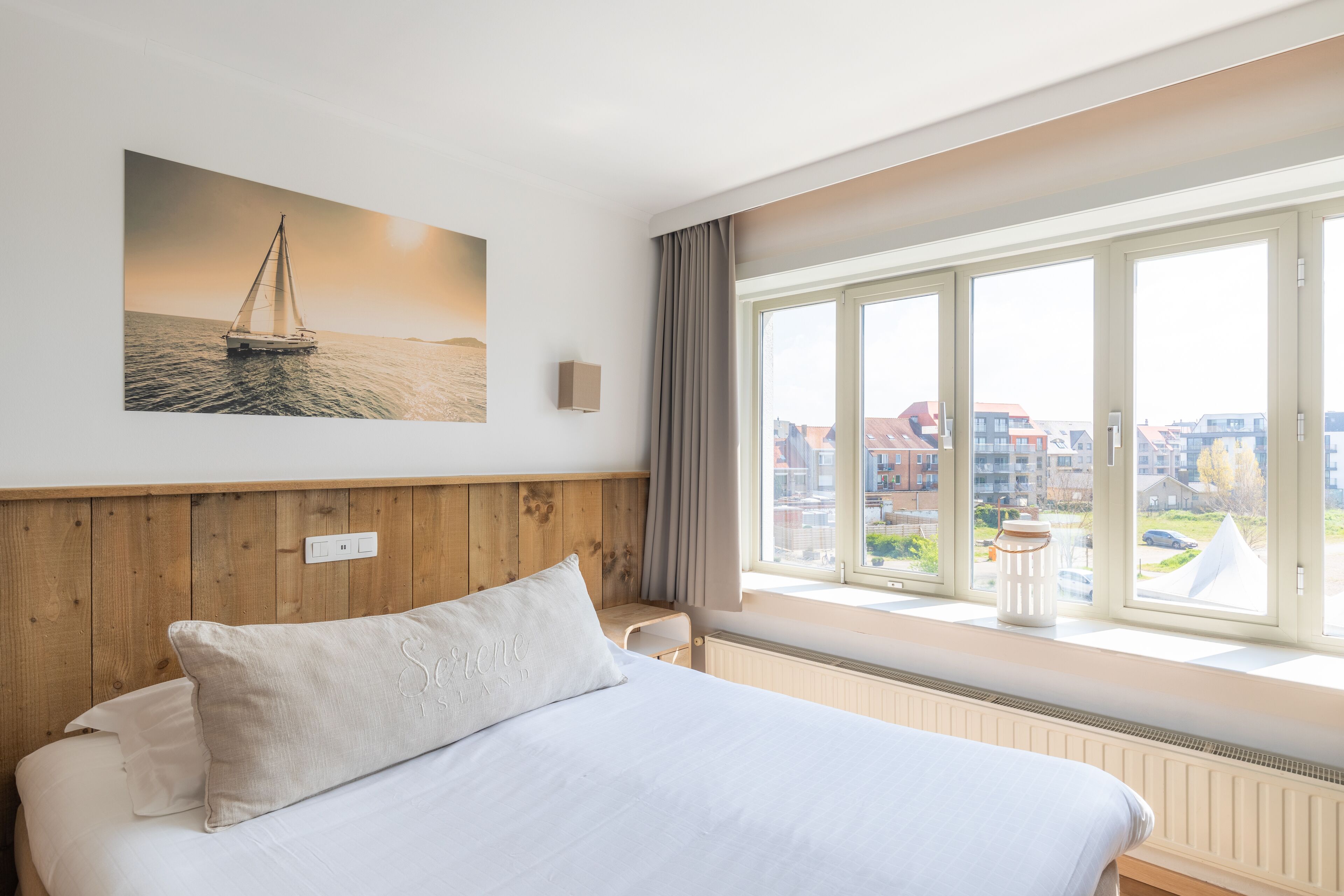 Habitació d'hotel amb un llit gran, vista de la ciutat a través de grans finestres i un quadre d'un veler a la paret.