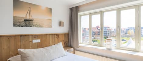 A hotel room with a large bed, a view of the city through large windows, and a painting of a sailboat on the wall.
