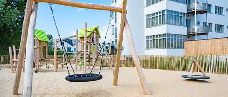 A playground with swings and a climbing structure, surrounded by a sandy area and a wooden fence.
