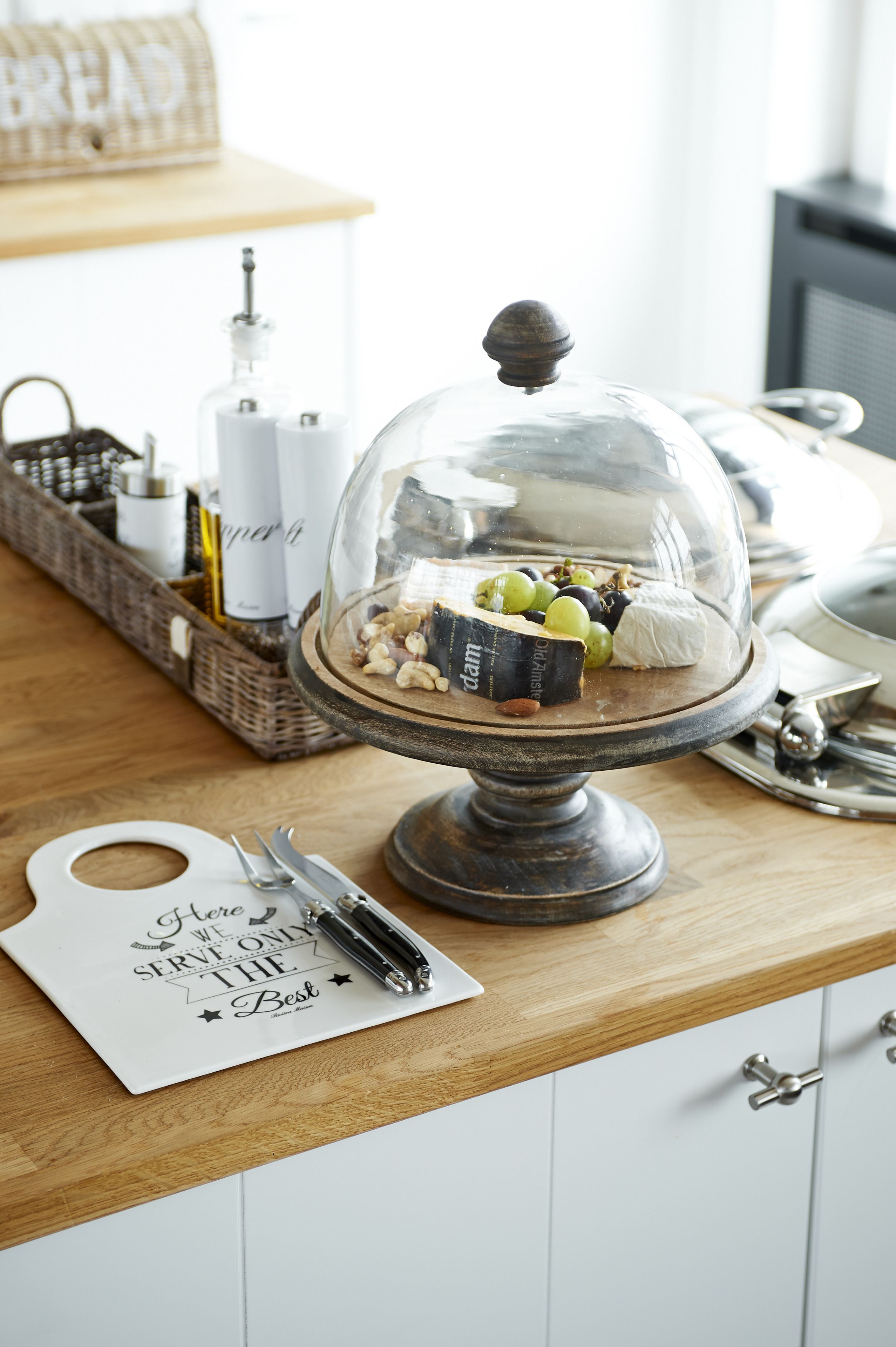 A wooden counter with a glass dome display case containing cheese, grapes, and nuts. A bottle of wine and a basket with bottles are visible in the background.