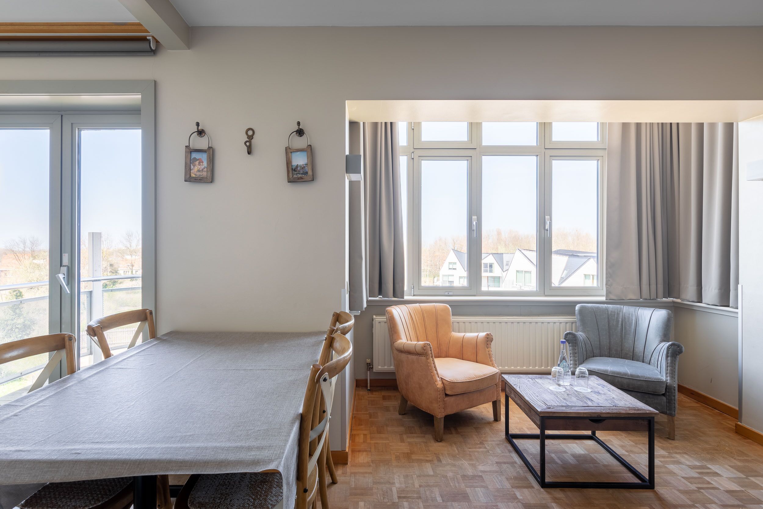 A spacious living room with a wooden dining table, two armchairs, a coffee table, and large windows with curtains.