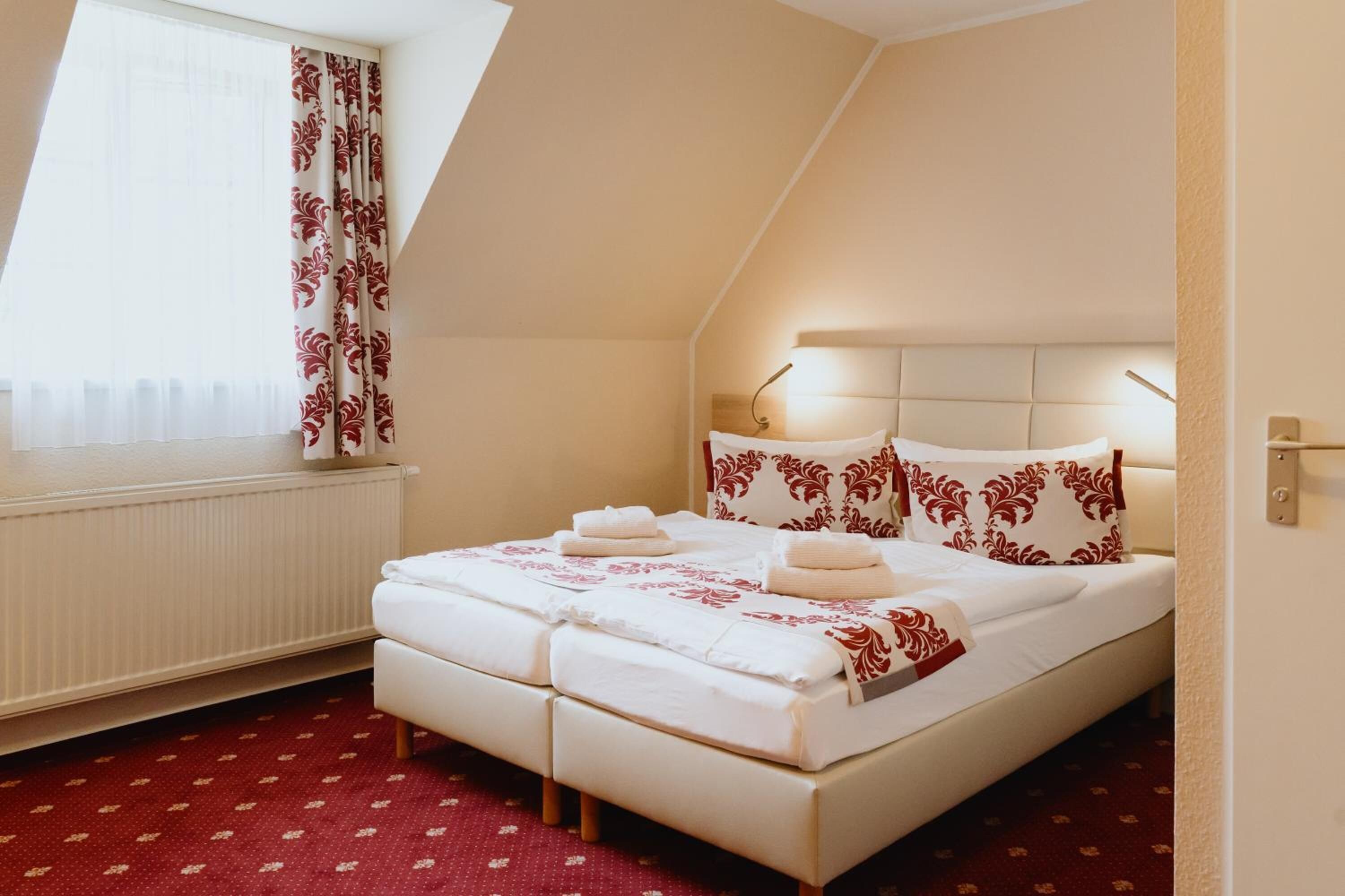 A neatly made bed with white linens and red floral patterns, a bedside lamp, and a window with sheer curtains.