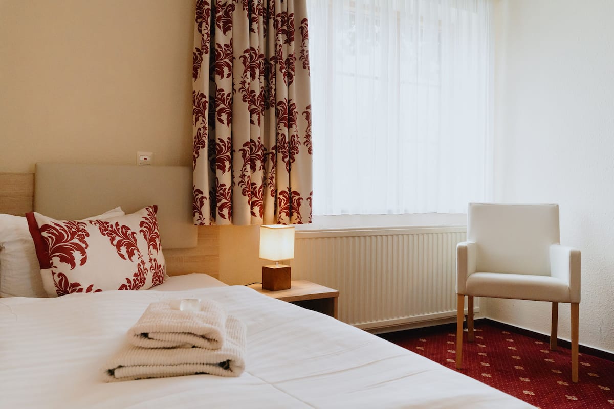 Una habitación de hotel con una cama bien hecha, una silla blanca, una mesita de noche con lámpara y un ventanal con cortinas.