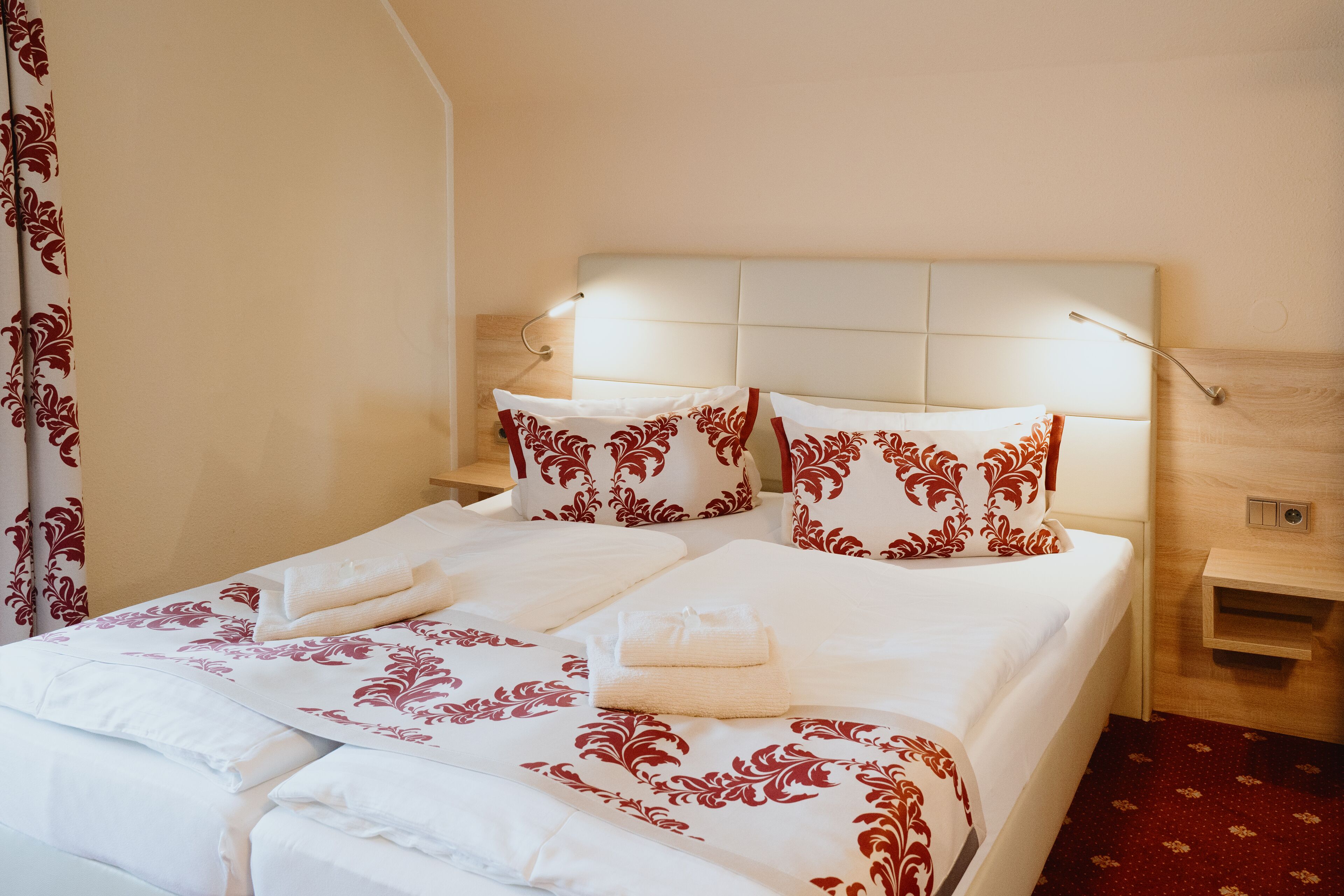 A neatly made bed with white and red floral bedding, a headboard with integrated lighting, and a wooden nightstand with a lamp.